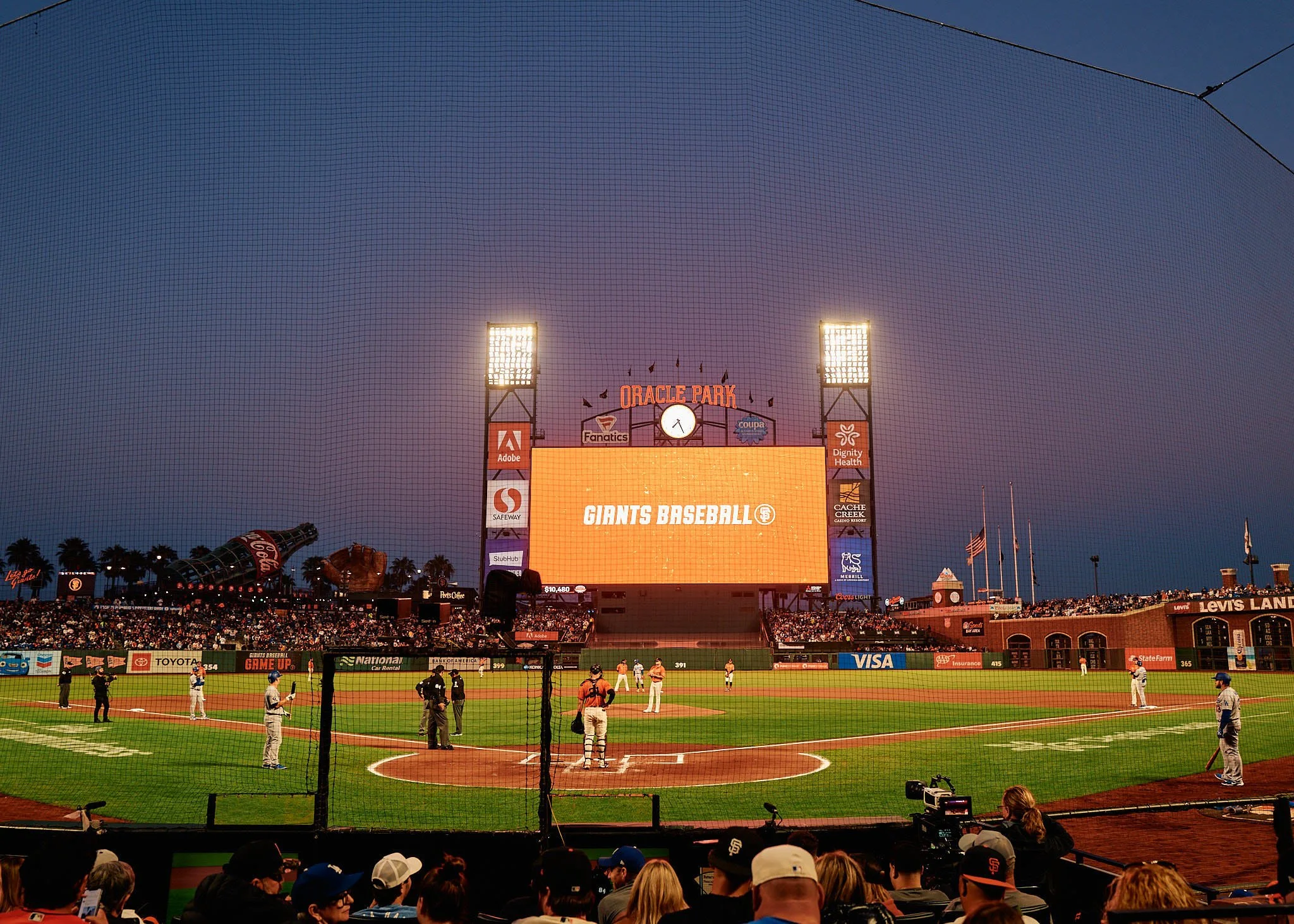 A baseball game at Oracle Park stadium during dusk, with players on the field and fans in the stands. The large scoreboard displays 'Giants Baseball' with surrounding advertisements.