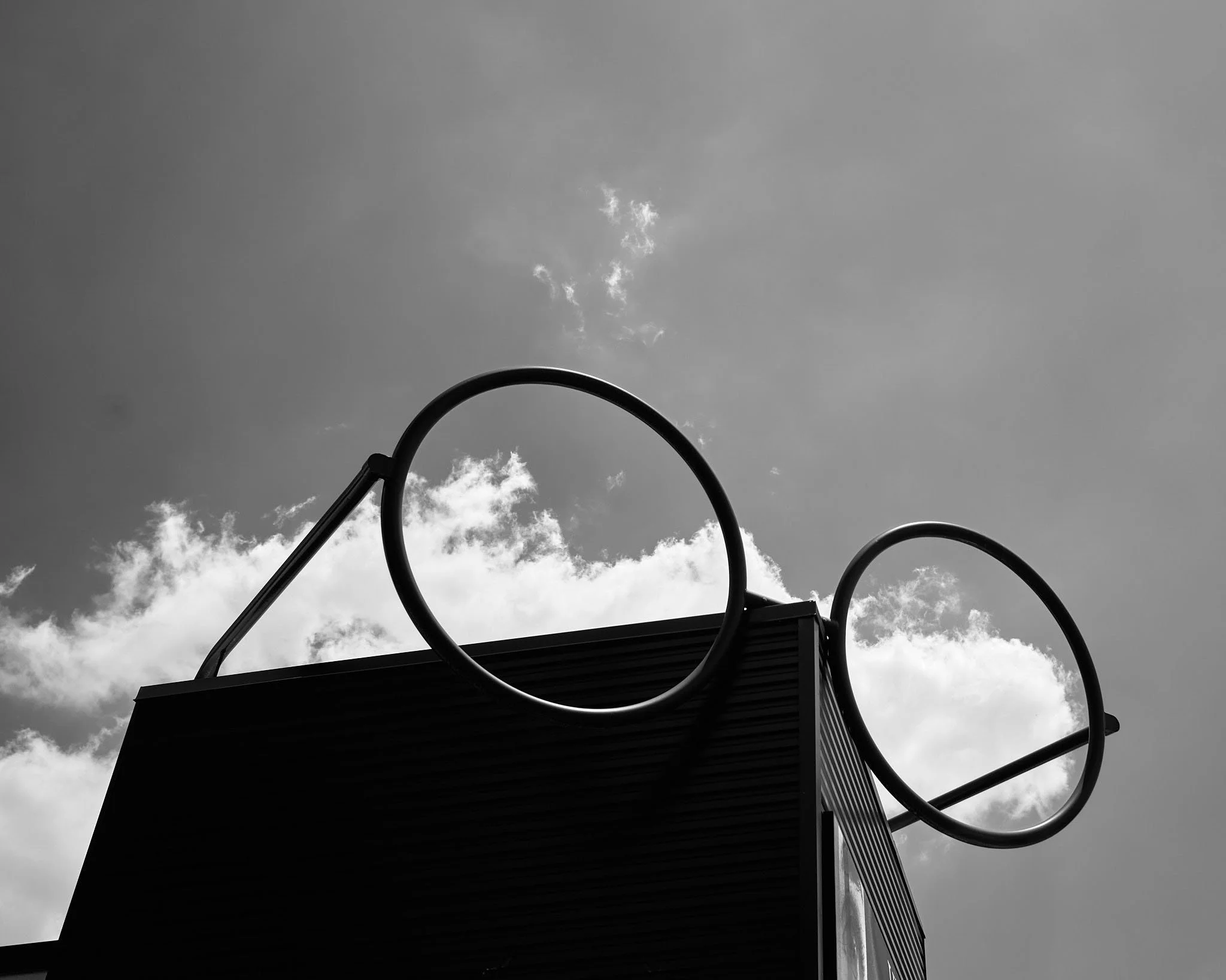Black and white photograph of a tall structure with two large circular hoops attached to the top, set against a cloudy sky.