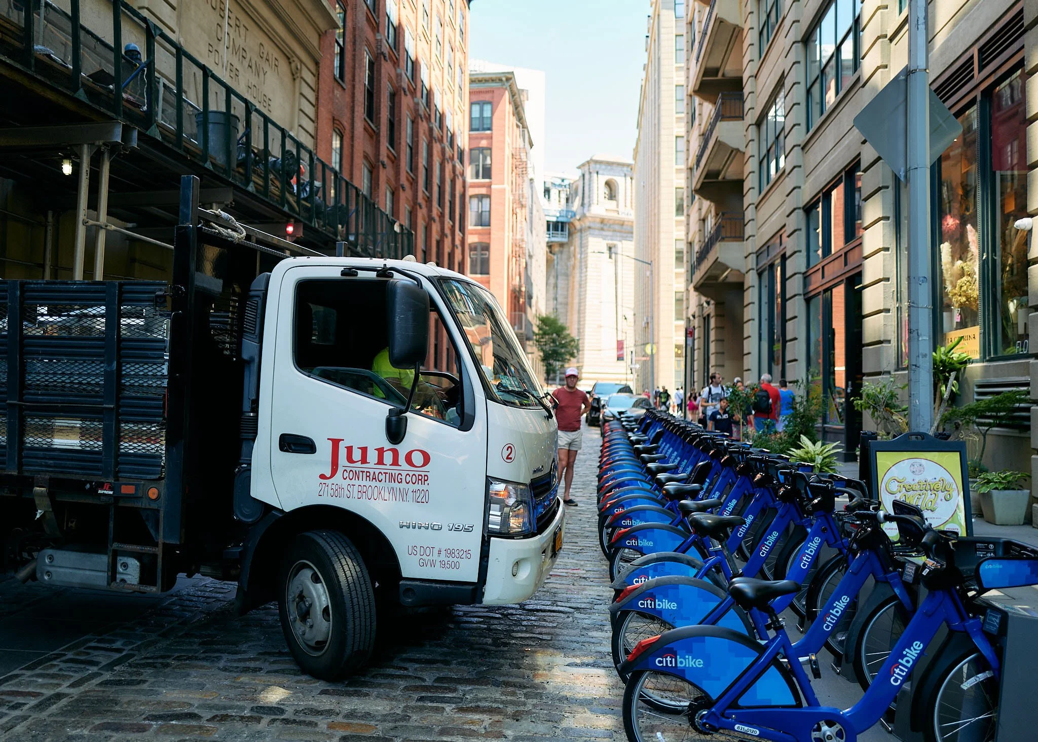 A city street scene with a white utility truck labeled Juno Contracting parked on a cobblestone street, and a row of blue CitiBike bicycles lined up along the sidewalk. Several pedestrians walk in the background, surrounded by tall brick and stone bu