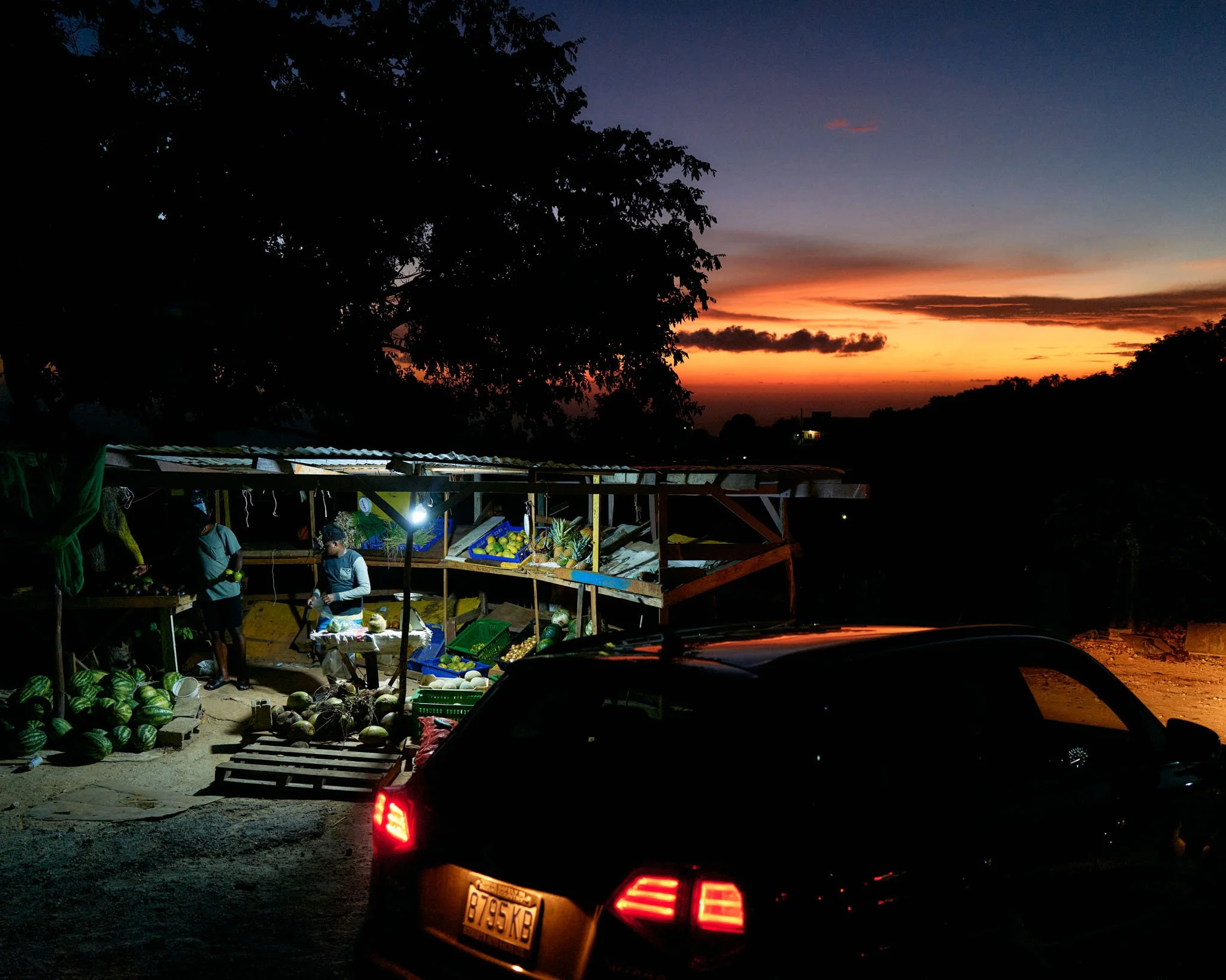 Nighttime outdoor market stall with produce and people, situated on dirt ground with a car parked in front. Sunset sky in the background with trees.