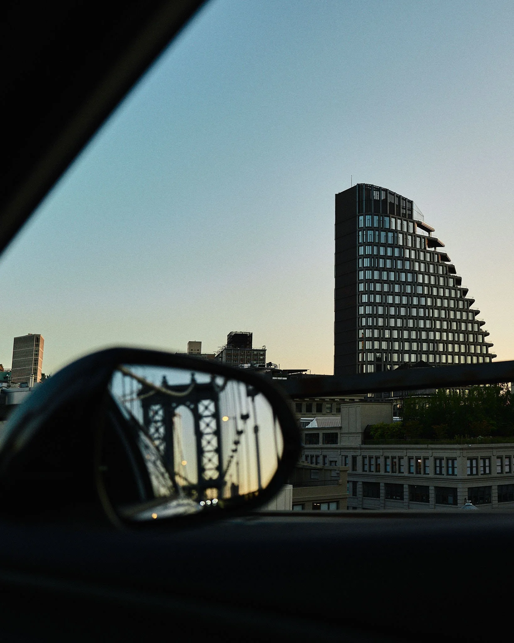 View of a city skyline at sunset with tall buildings, including a prominent modern high-rise, seen through the rearview mirror of a car. The car's window and mirror frame the scene.