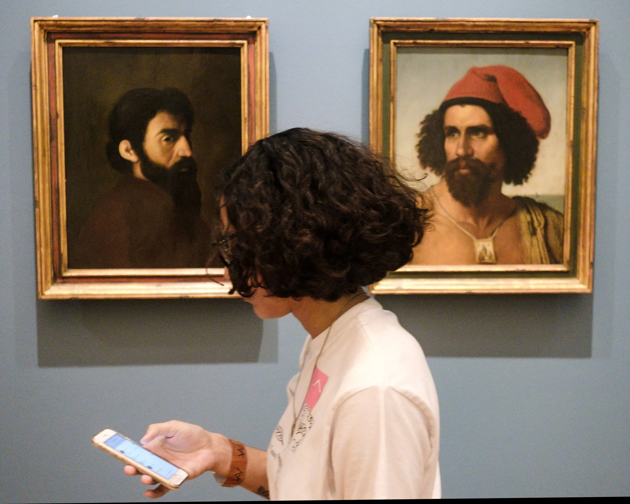 Person with curly hair using a smartphone in front of two framed portraits of men with beards.