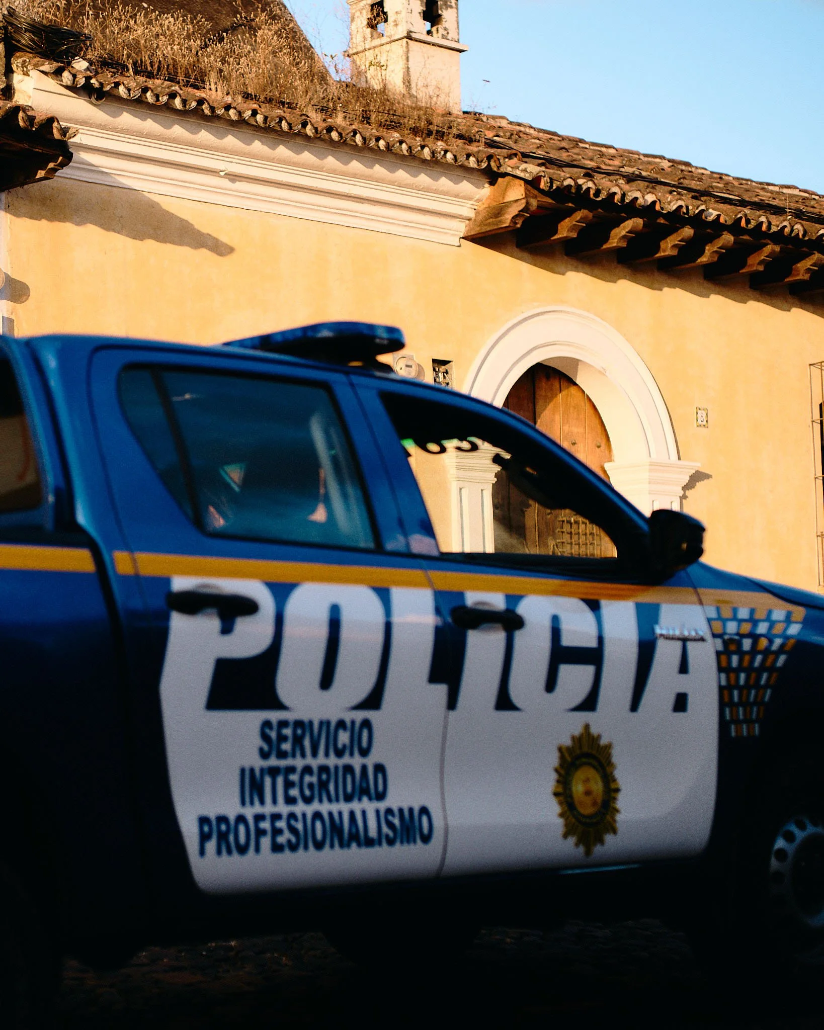 A Mexican police car with the words 'Policía Servicio Integridad Profesionalismo' on the side, parked in front of a yellow building with a wooden arched door and a bell tower in the background.