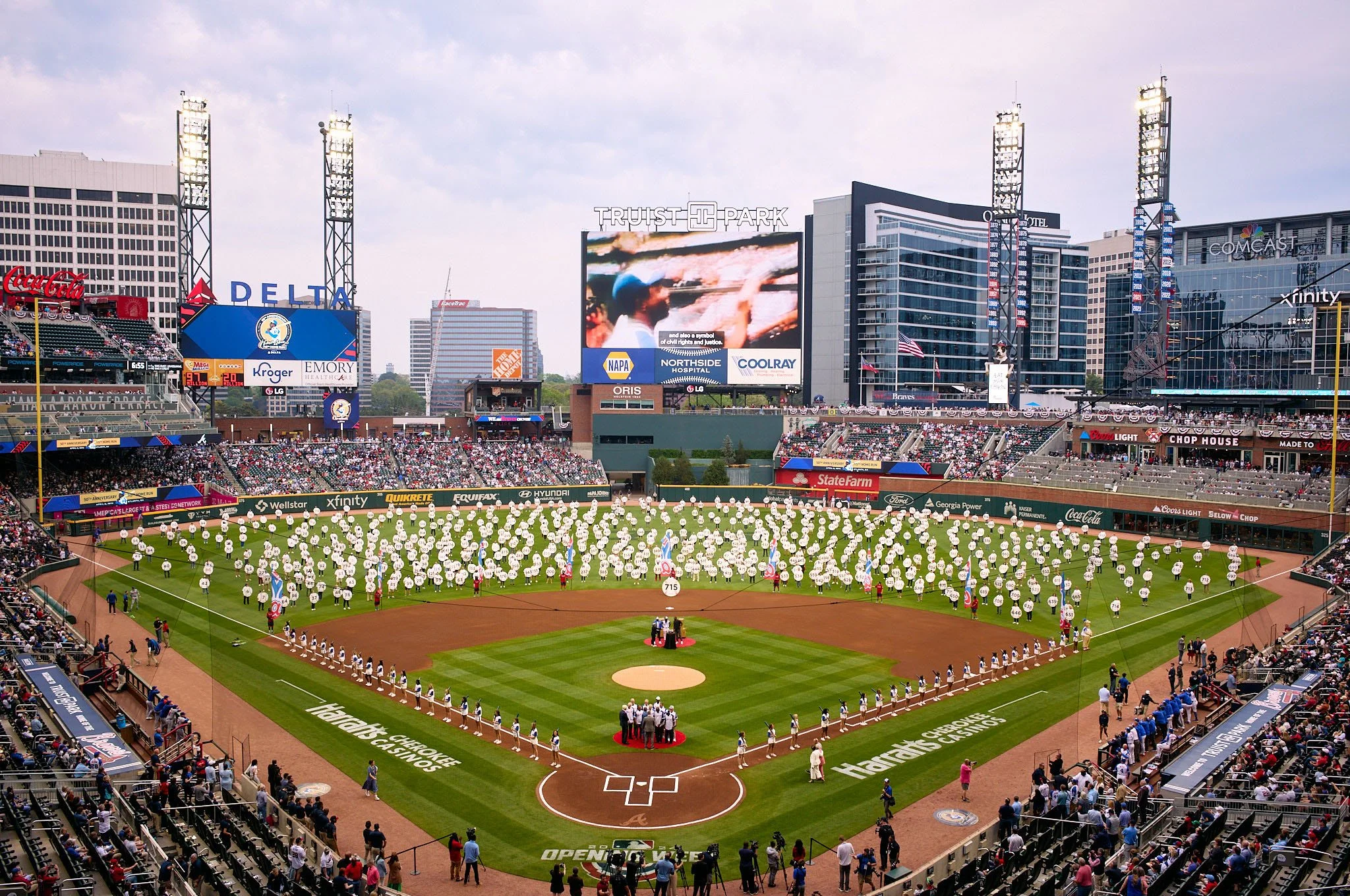 People at a baseball stadium holding white balloons while a ceremony is taking place on the field. The stadium has a large digital scoreboard and surrounding skyscrapers.