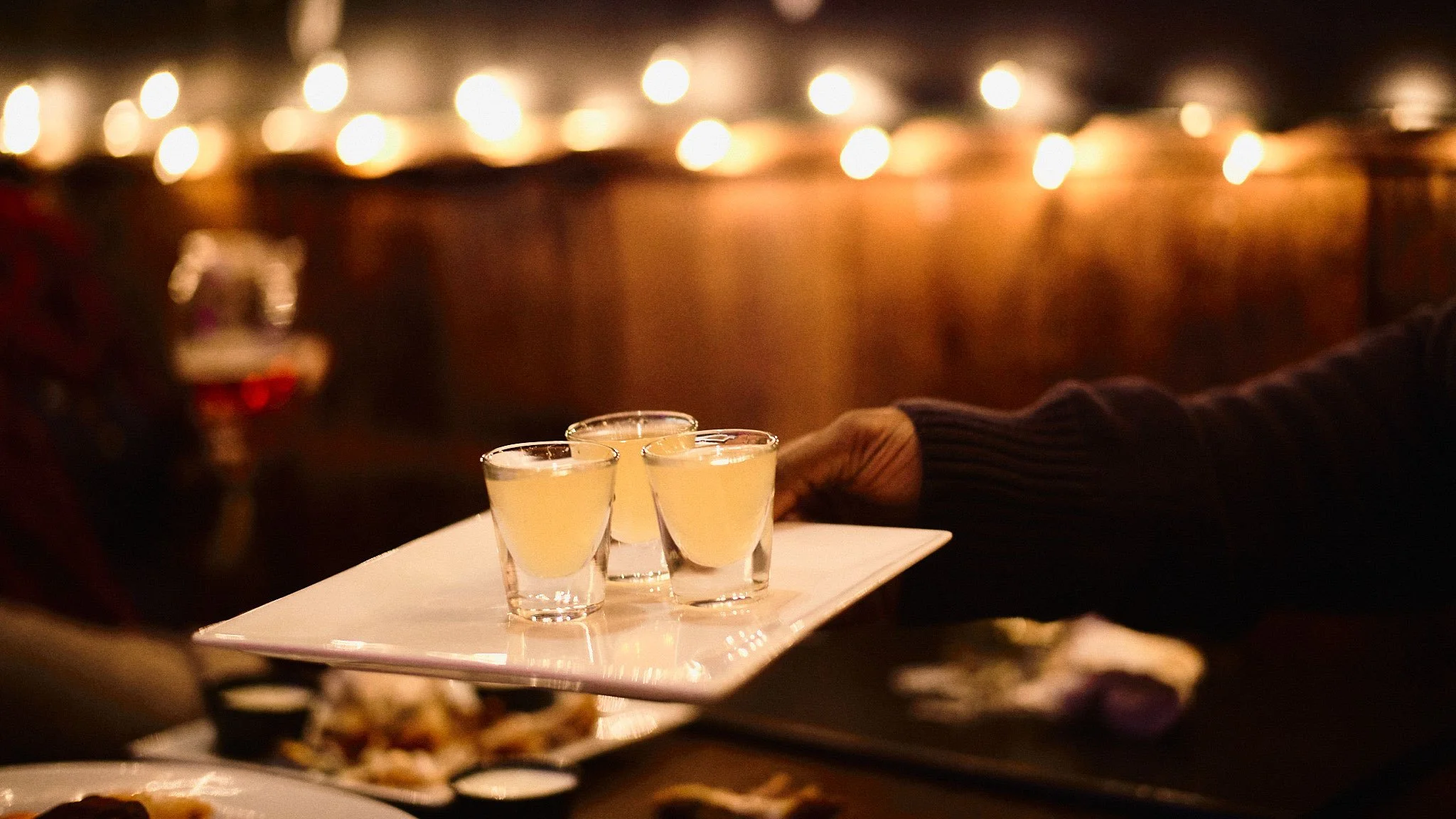 A person holding a tray with three shot glasses filled with yellowish drinks at a dimly lit restaurant or bar.