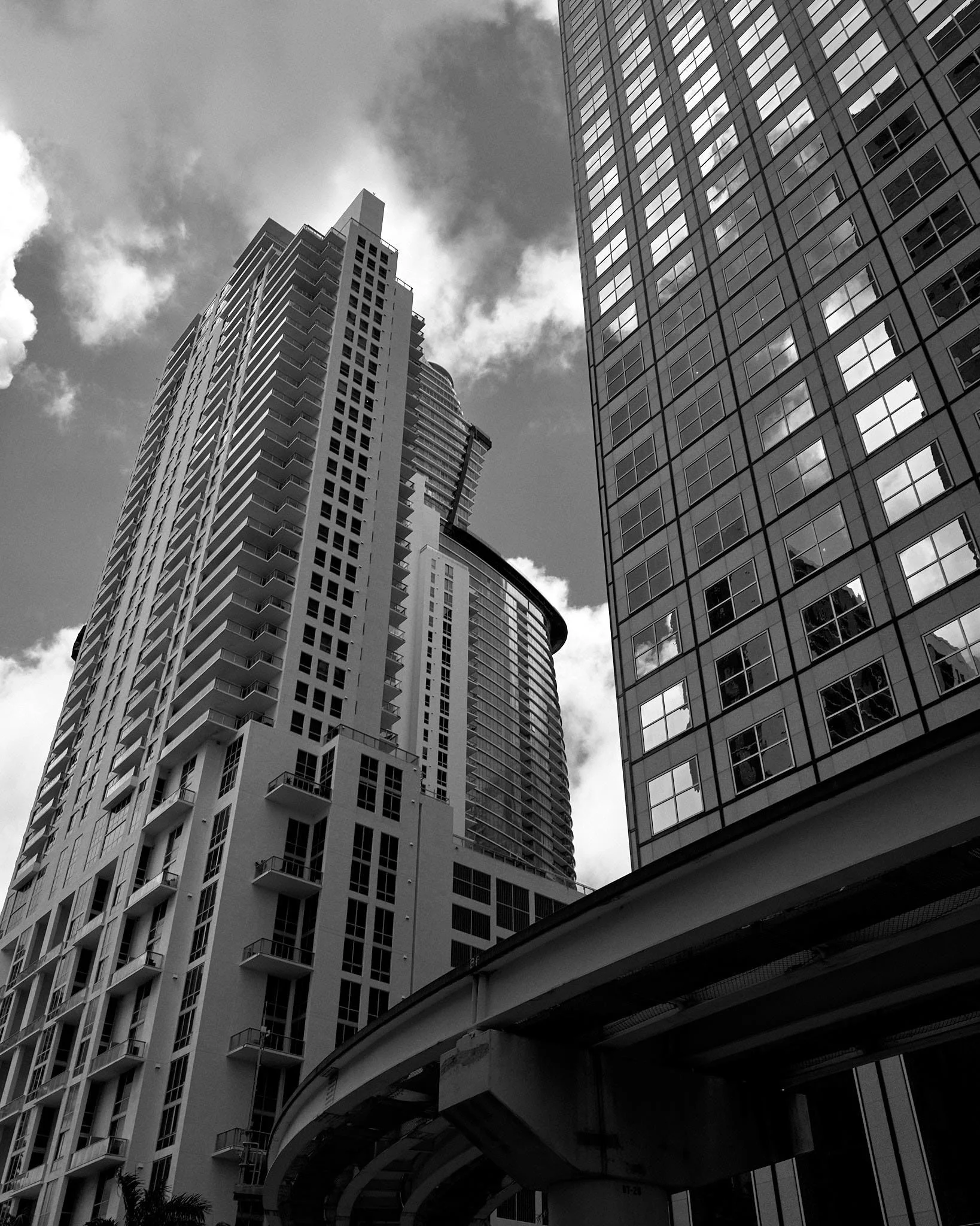 Black and white photo of tall modern skyscrapers with many windows, some reflecting cloudy sky, taken from below.