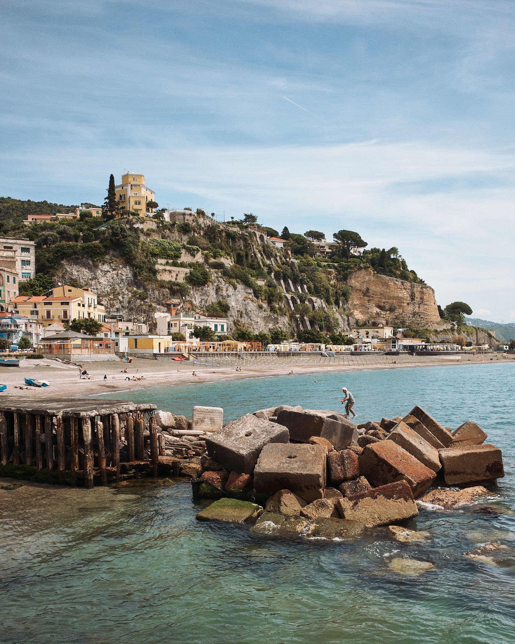 Coastal scene with a rocky shoreline, calm water, a man fishing on rocks, beach with people, hillside with houses and a yellow building, and a partly cloudy sky.