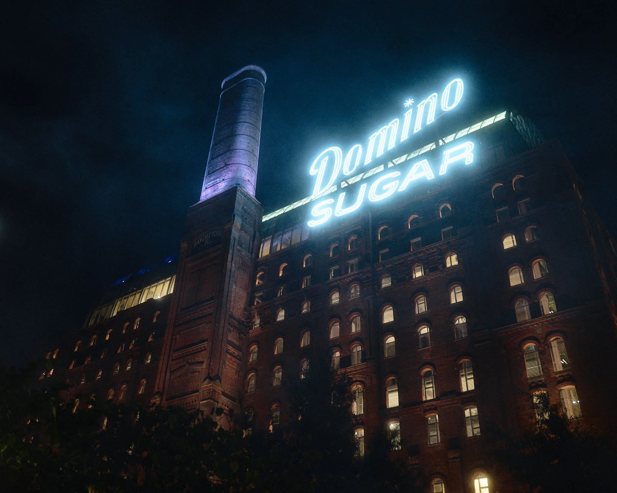Night view of a large brick building with a neon sign reading 'Domino Sugar' and an illuminated chimney.