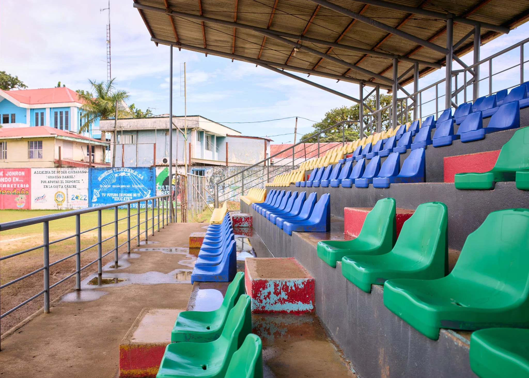 Empty stadium stands with brightly colored green, blue, and yellow seats on a muddy field under a temporary roof. Residential buildings and advertisements are visible in the background.