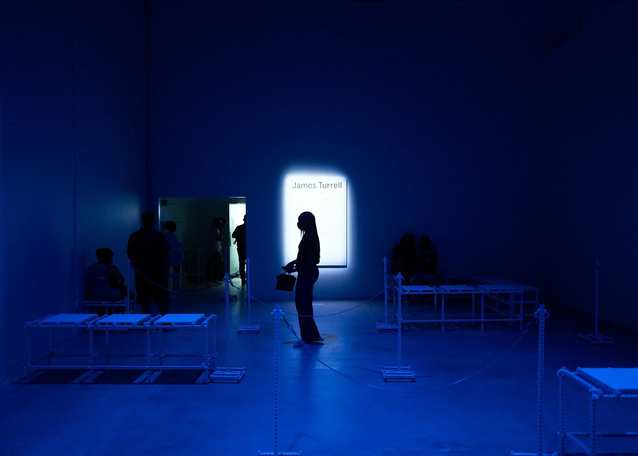 An art gallery with dim blue lighting, a lit display with the name 'James Turrell' on the wall, and several visitors viewing the exhibit from a distance.