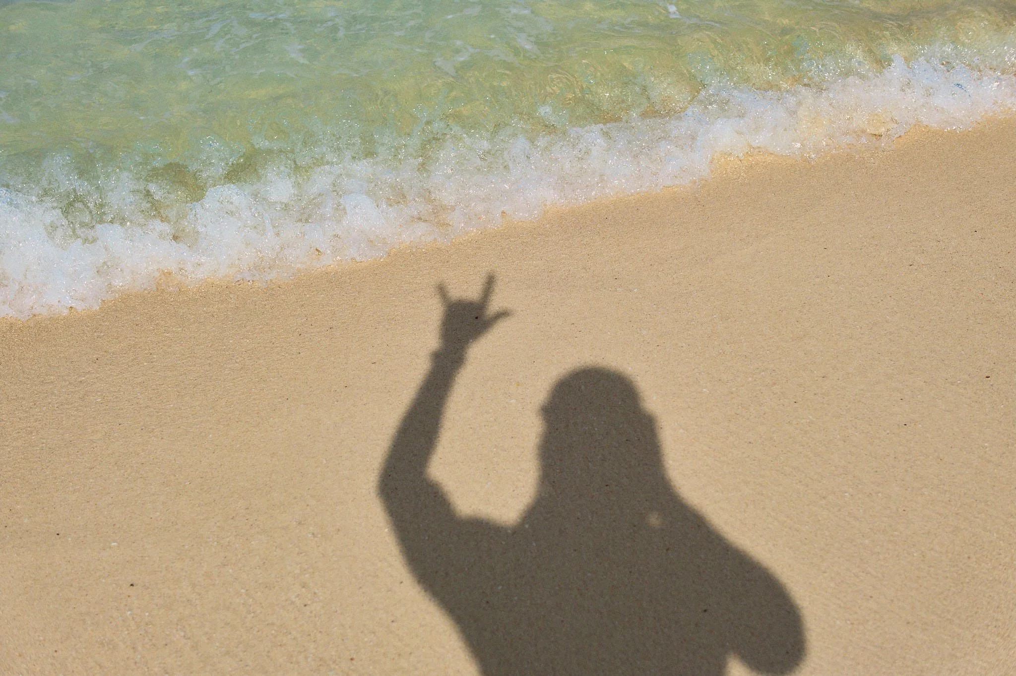 Shadow of a person on the sandy beach making a peace sign with the hand, with the ocean and small waves in the background.