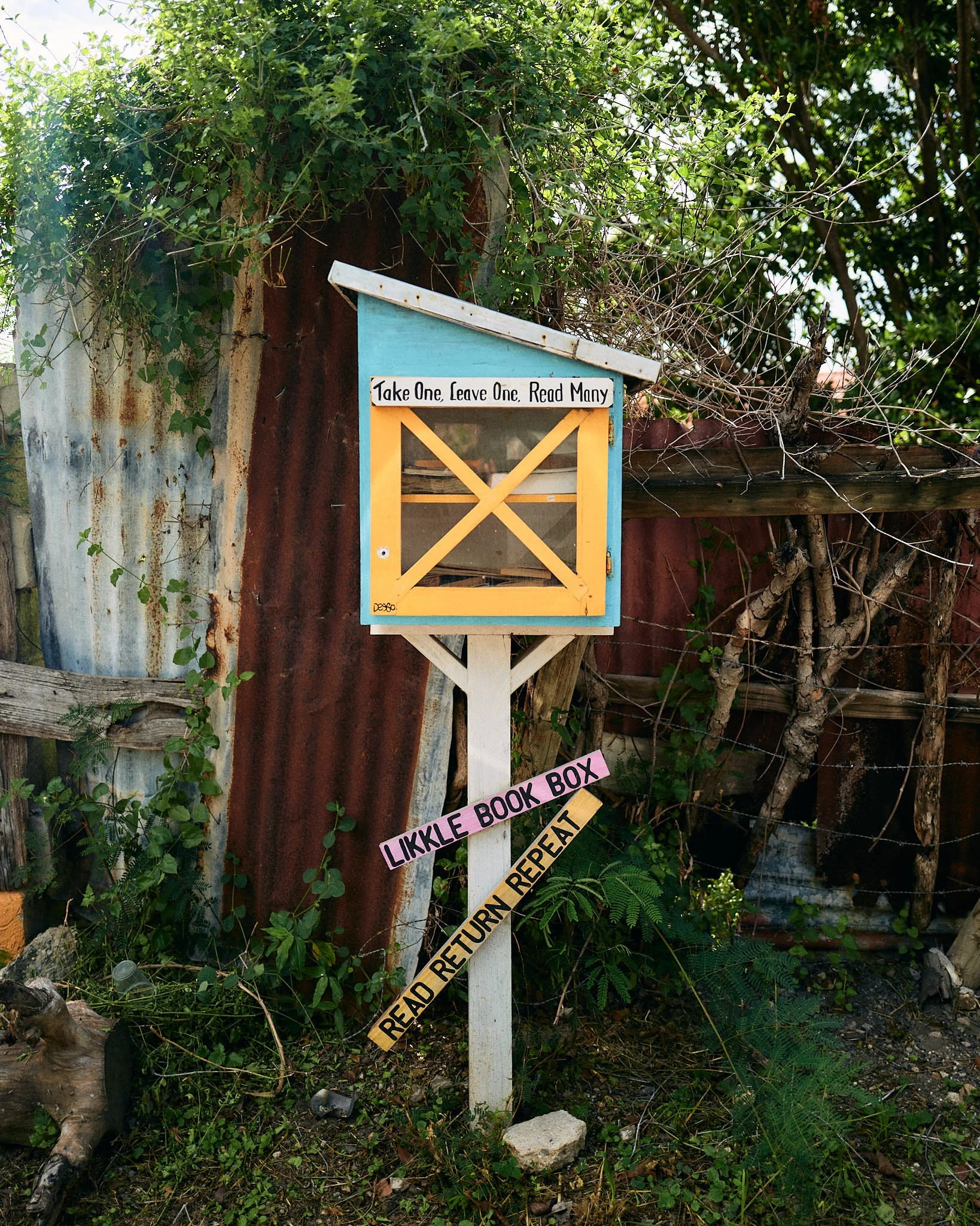 A small, colorful outdoor book exchange box on a post in a garden or yard, with a sign saying "Take One Leave One Read Many" and another sign below saying "Likkle Book Box" and "Read Return Repeat".
