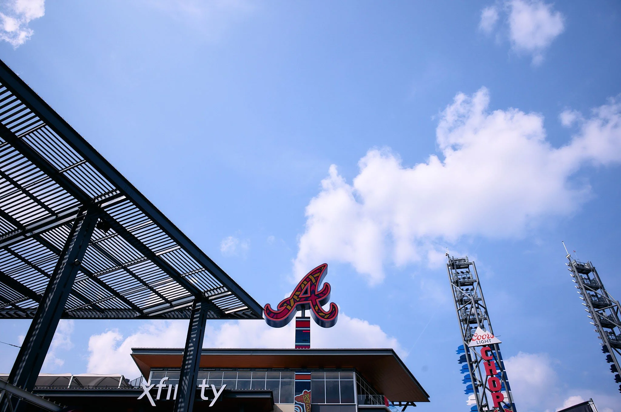 View of the Atlanta Braves stadium featuring the team’s logo, a roof structure on the left, and tall towers with signs, all set against a blue sky with clouds.