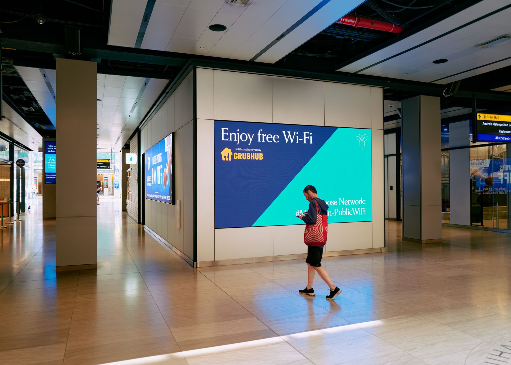 A man in a red backpack walks through an airport terminal, looking at his phone. Behind him is a digital sign advertising free Wi-Fi sponsored by Grubhub.