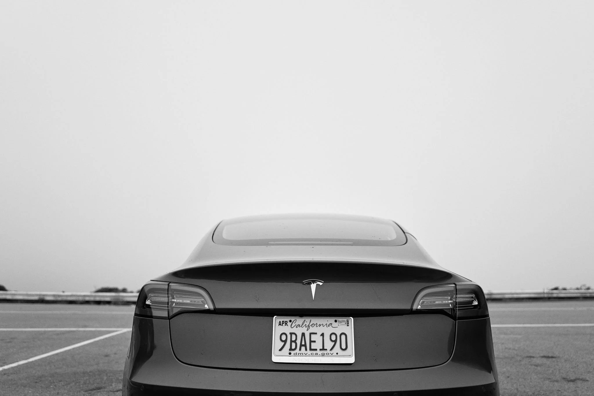 Black Tesla Model S parked in an empty lot, viewed from the rear, with a California license plate reading 9BAE190.