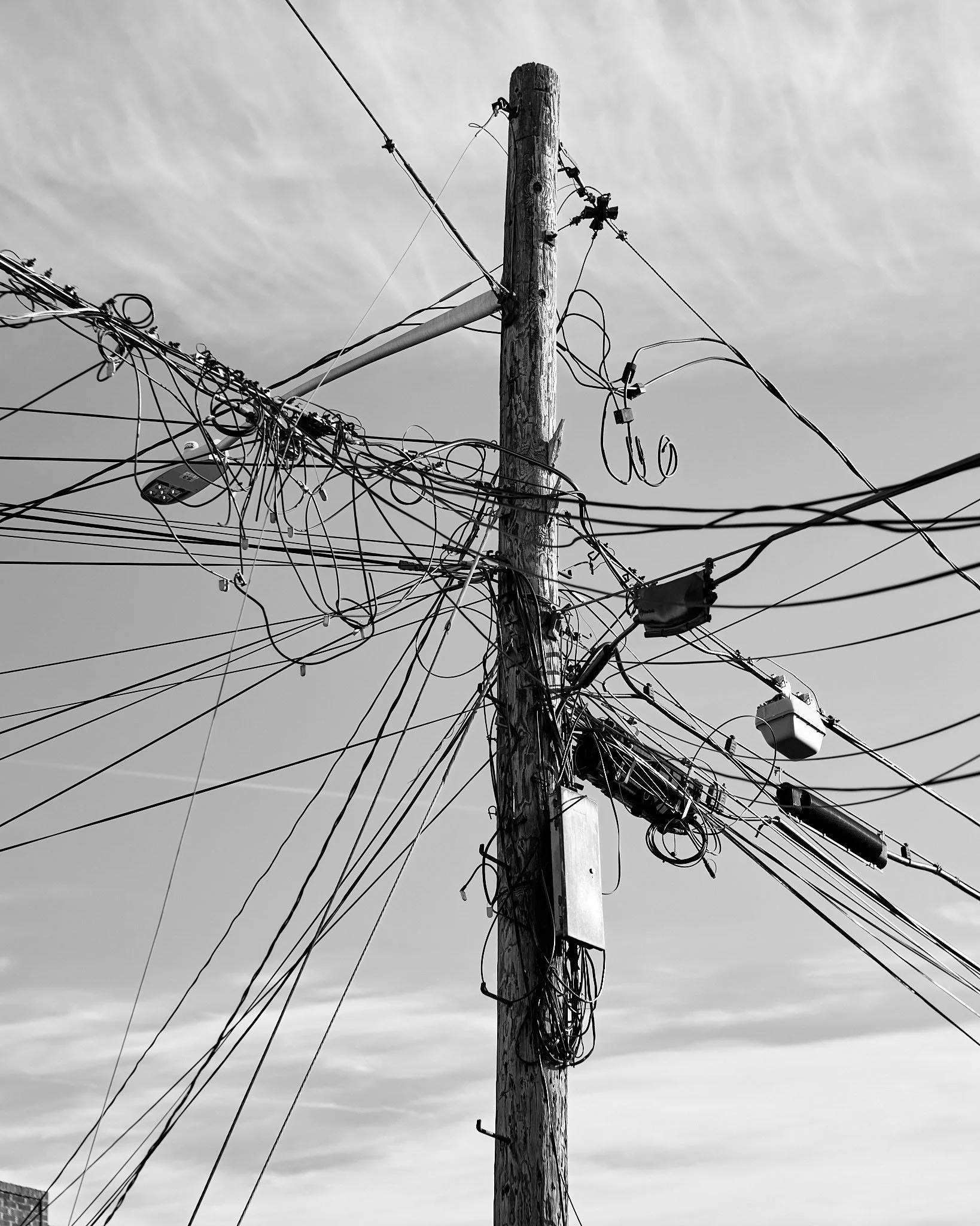 A utility pole with many tangled wires and electrical components against a cloudy sky in black and white.