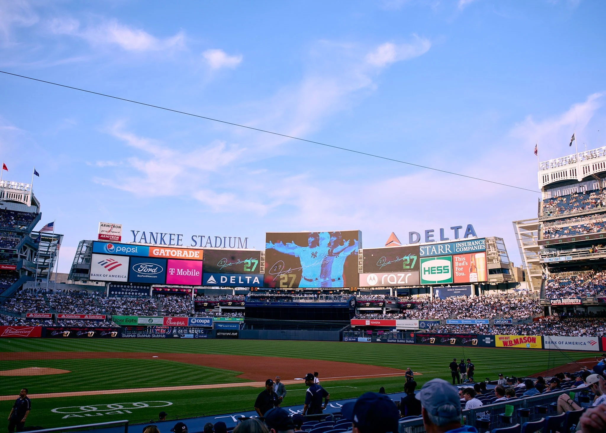 View of Yankee Stadium during a baseball game, with a large scoreboard displaying players and advertisements, and fans seated in the stands under a blue sky with some clouds.