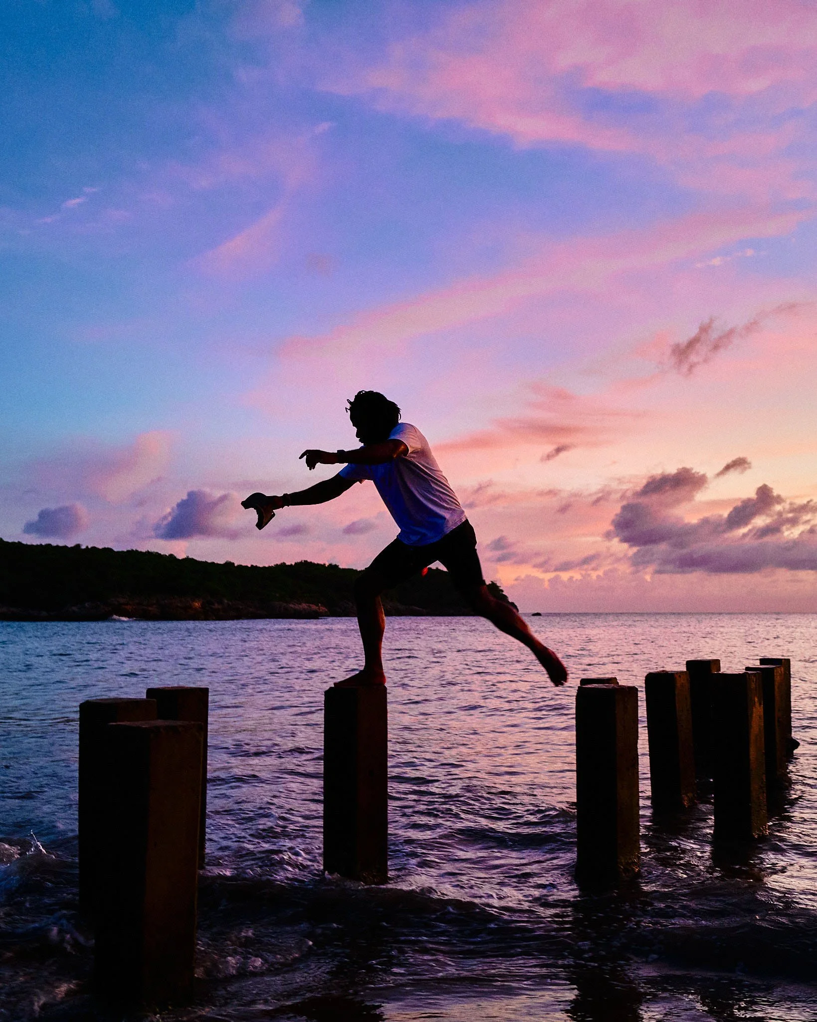 A person balancing on wooden posts in the water during a colorful sunset.