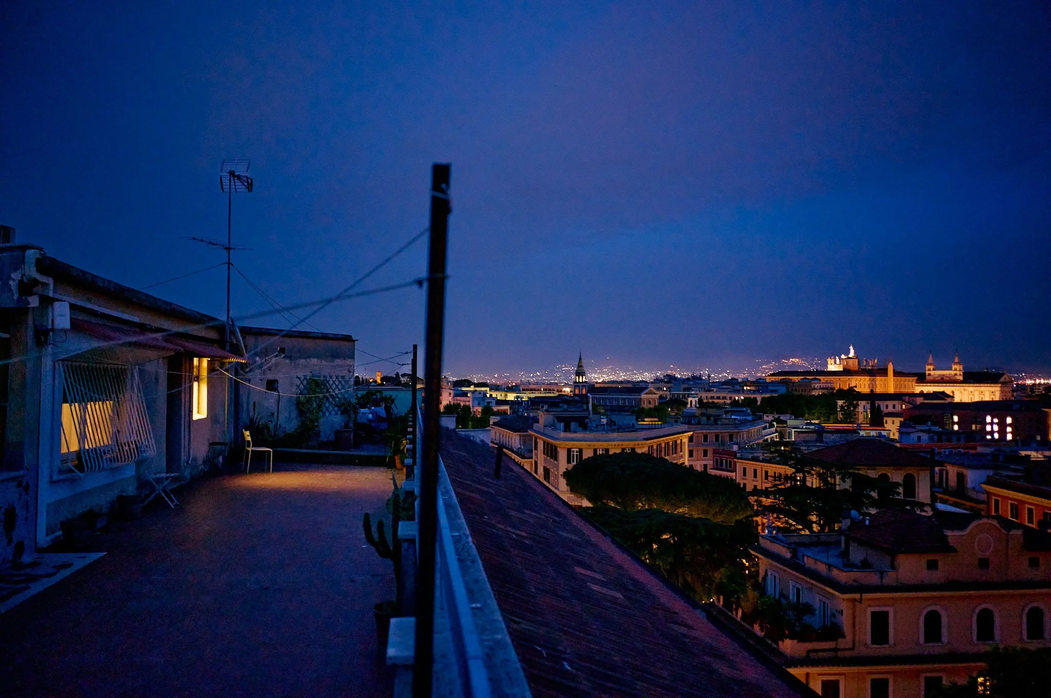 Nighttime cityscape view from a rooftop terrace with string lights, buildings, and lit landmarks in the distance.