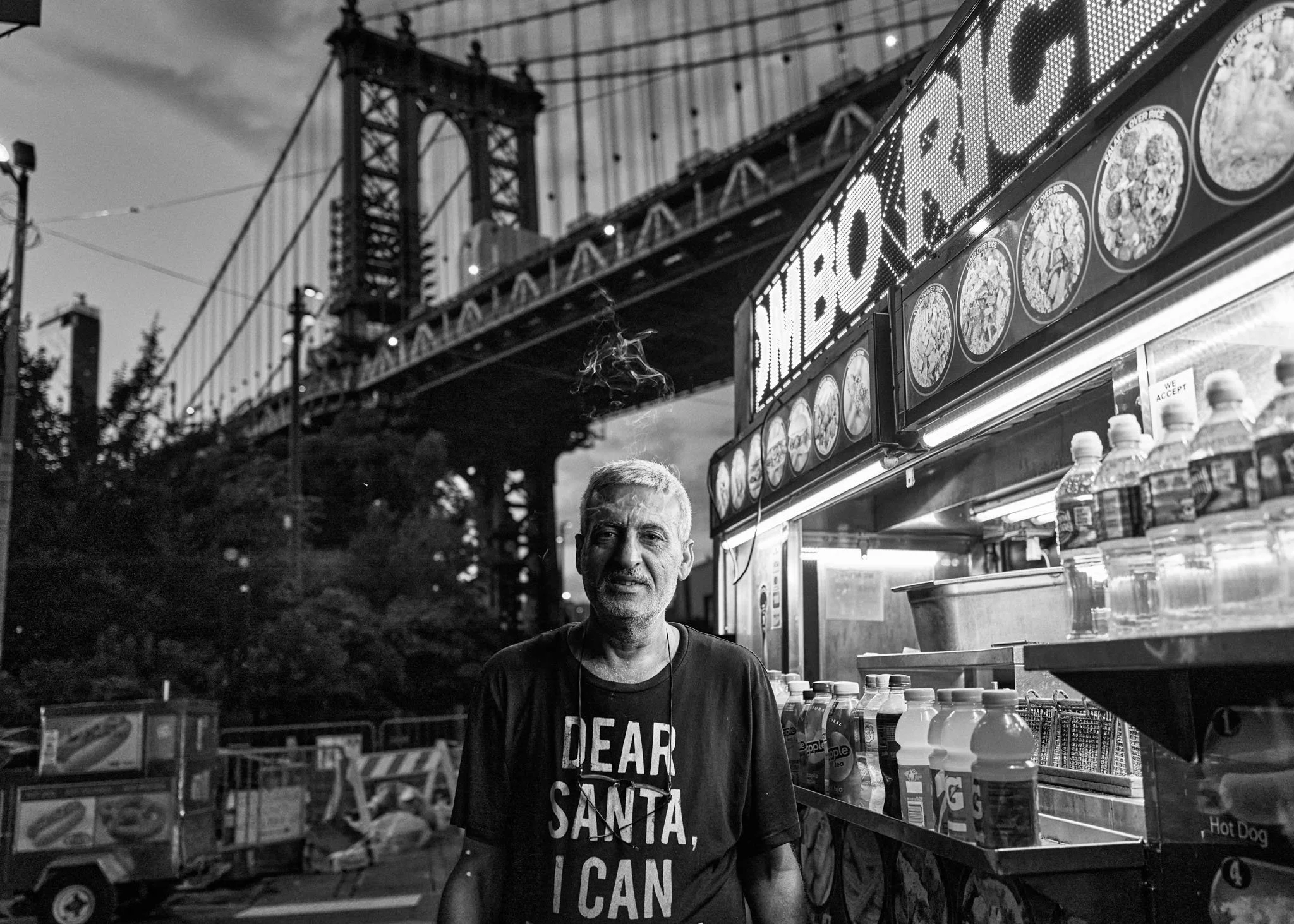 A man with a beard and short gray hair standing at a hot dog stand near the Brooklyn Bridge in New York City, black and white photo.