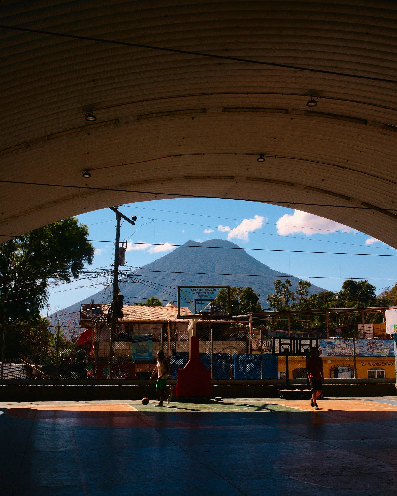 Children playing basketball on an outdoor court with a mountain in the background, viewed from under a curved roof.