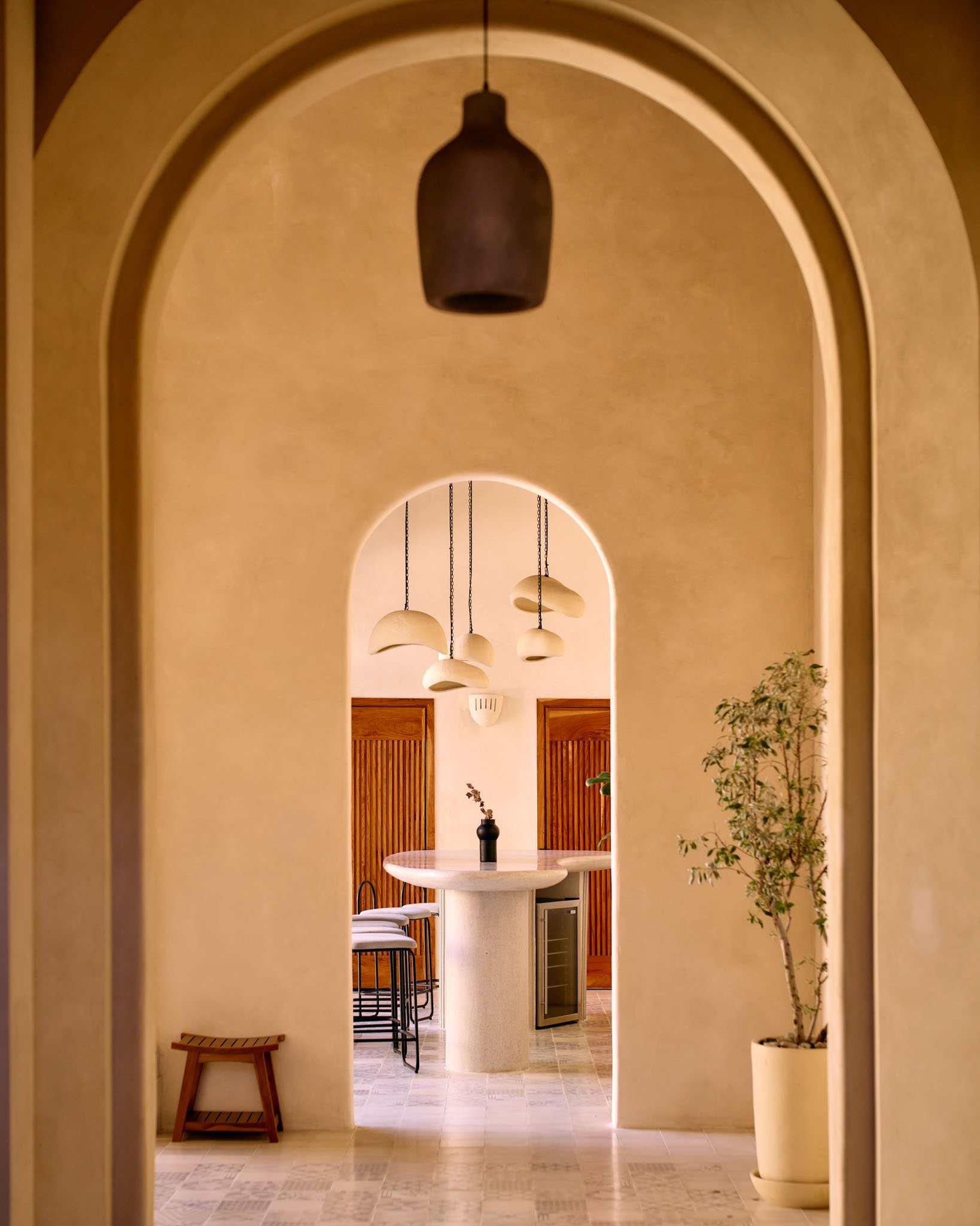 A view through a series of open archways into a modern, minimalist interior with a white round table, hanging pendant lights, and wooden doors.