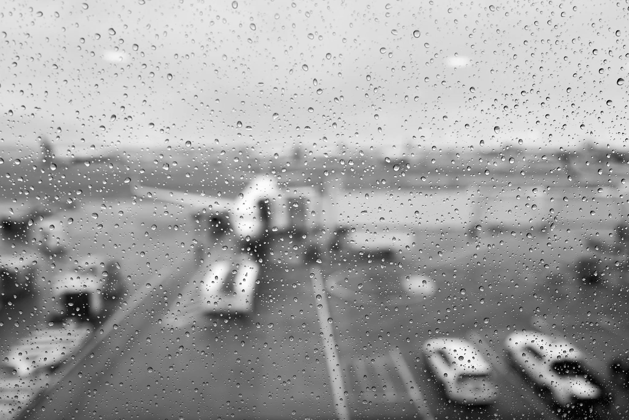 Rainy view from an airplane window with droplets on the glass, airport tarmac with parked airplanes and cars visible in the background.