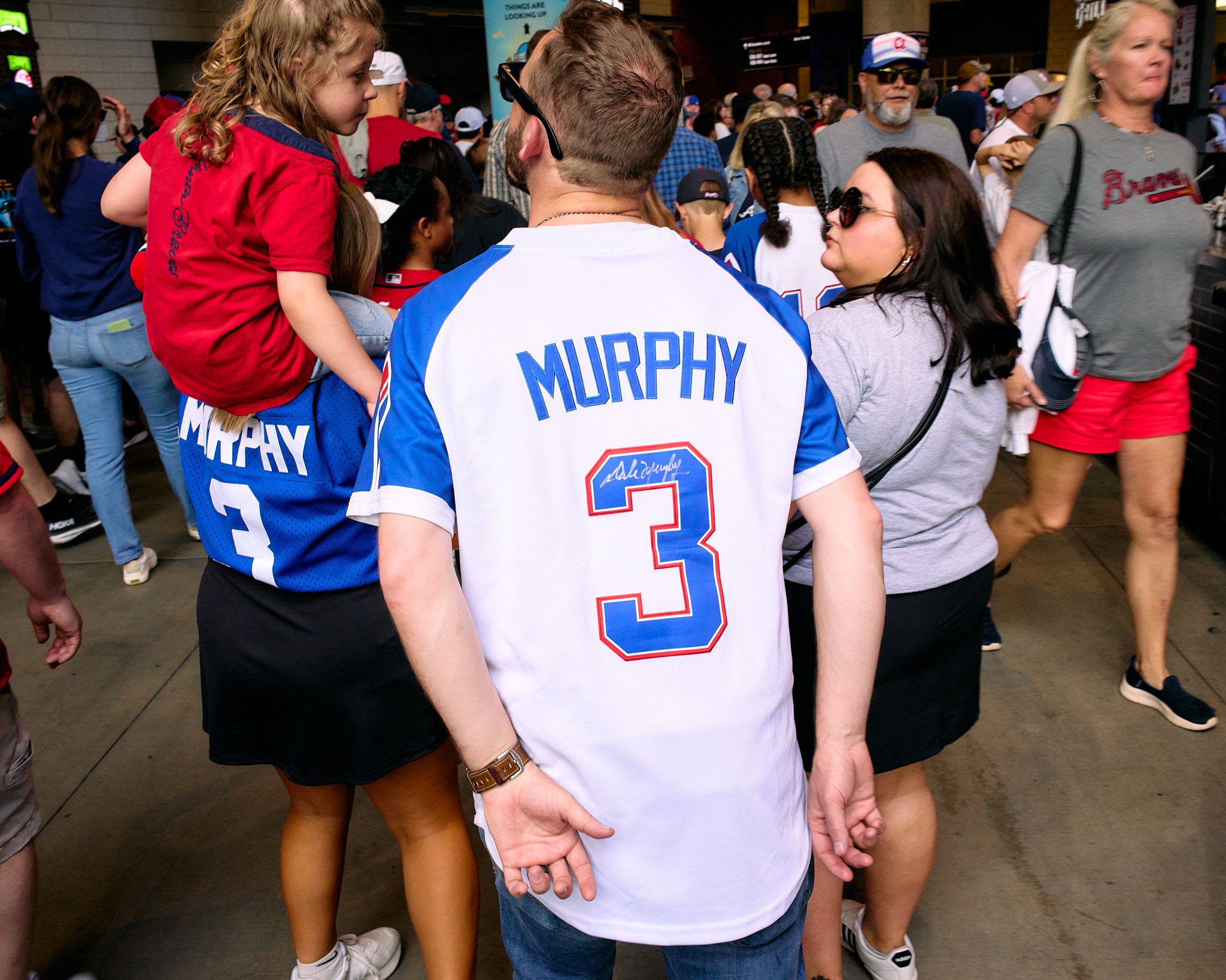 A crowd of sports fans, many wearing Atlanta Braves apparel, gathered at a stadium entrance. A man in the center wearing a white and blue Atlanta Braves jersey with the name 'Murphy' and the number 3 on it is visible from behind. A woman standing nex