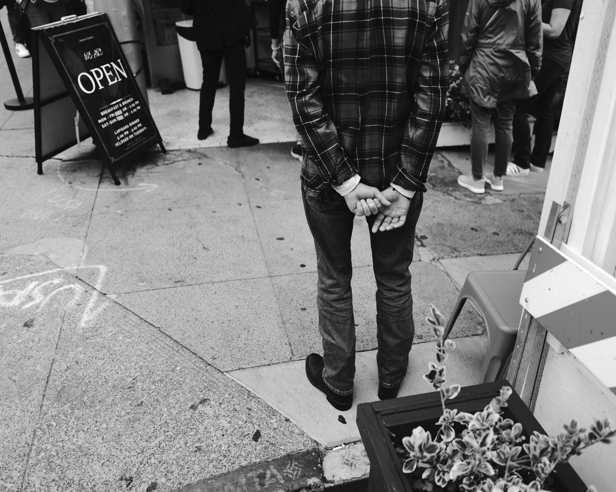 A person standing on a sidewalk near a restaurant entrance, with their hands clasped behind their back. The scene is in black and white, with a sign indicating the restaurant is open and a group of people waiting in line.