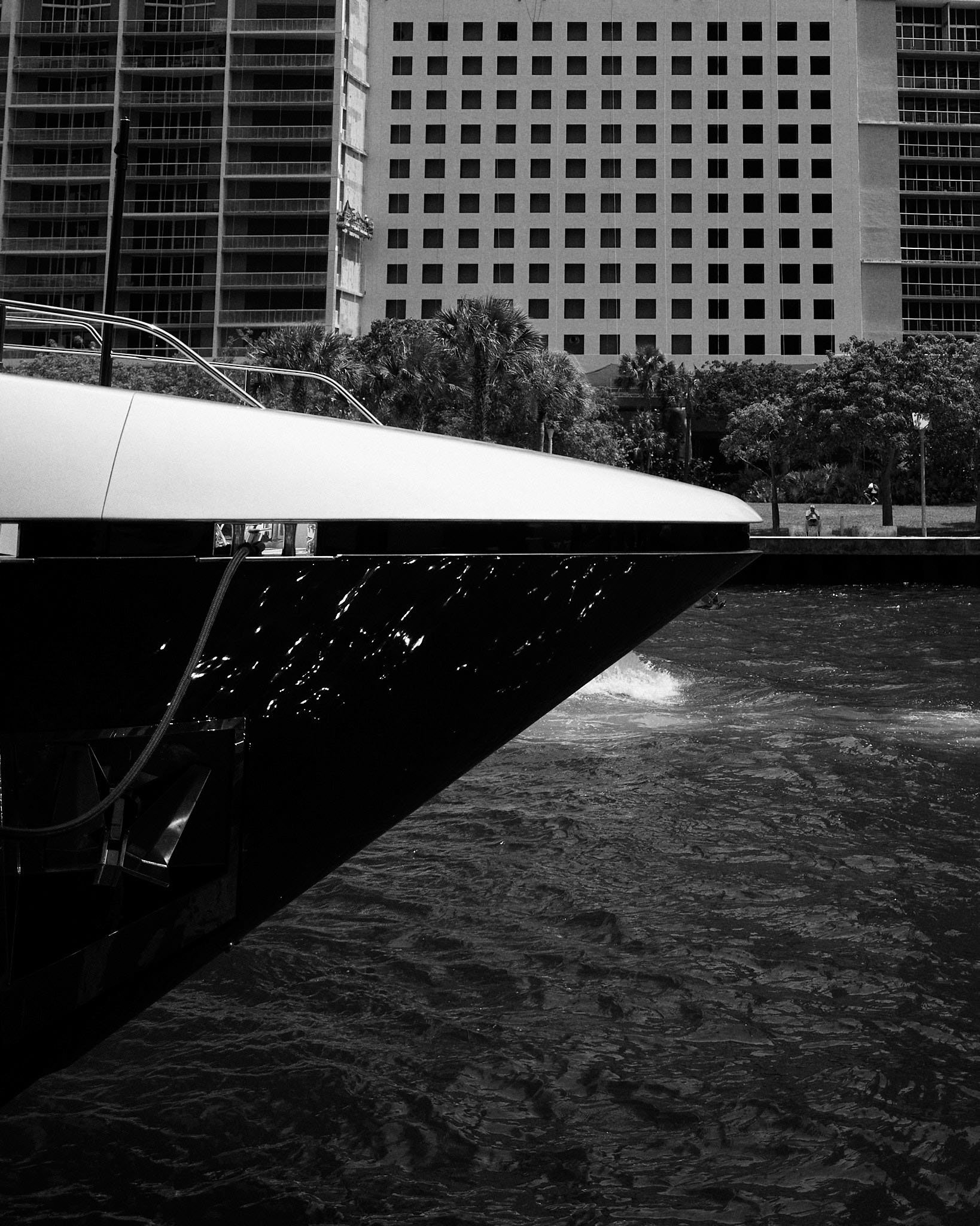Close-up of a yacht's bow on water with city buildings and trees in the background, black and white photograph.