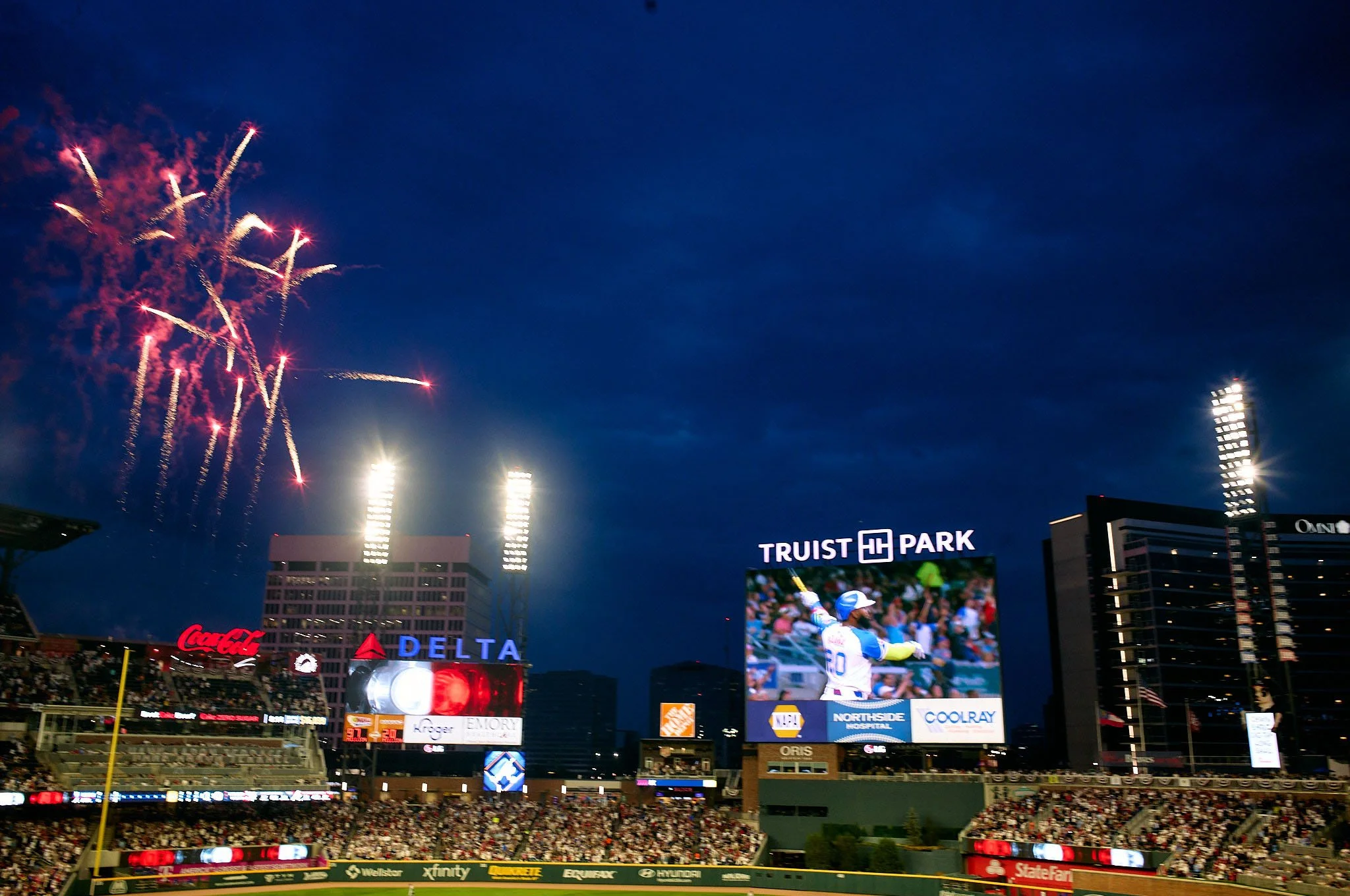 Fireworks in the night sky above Truist Park stadium with a baseball game on the large video screen. The stadium is filled with spectators, and bright lights illuminate the scene.