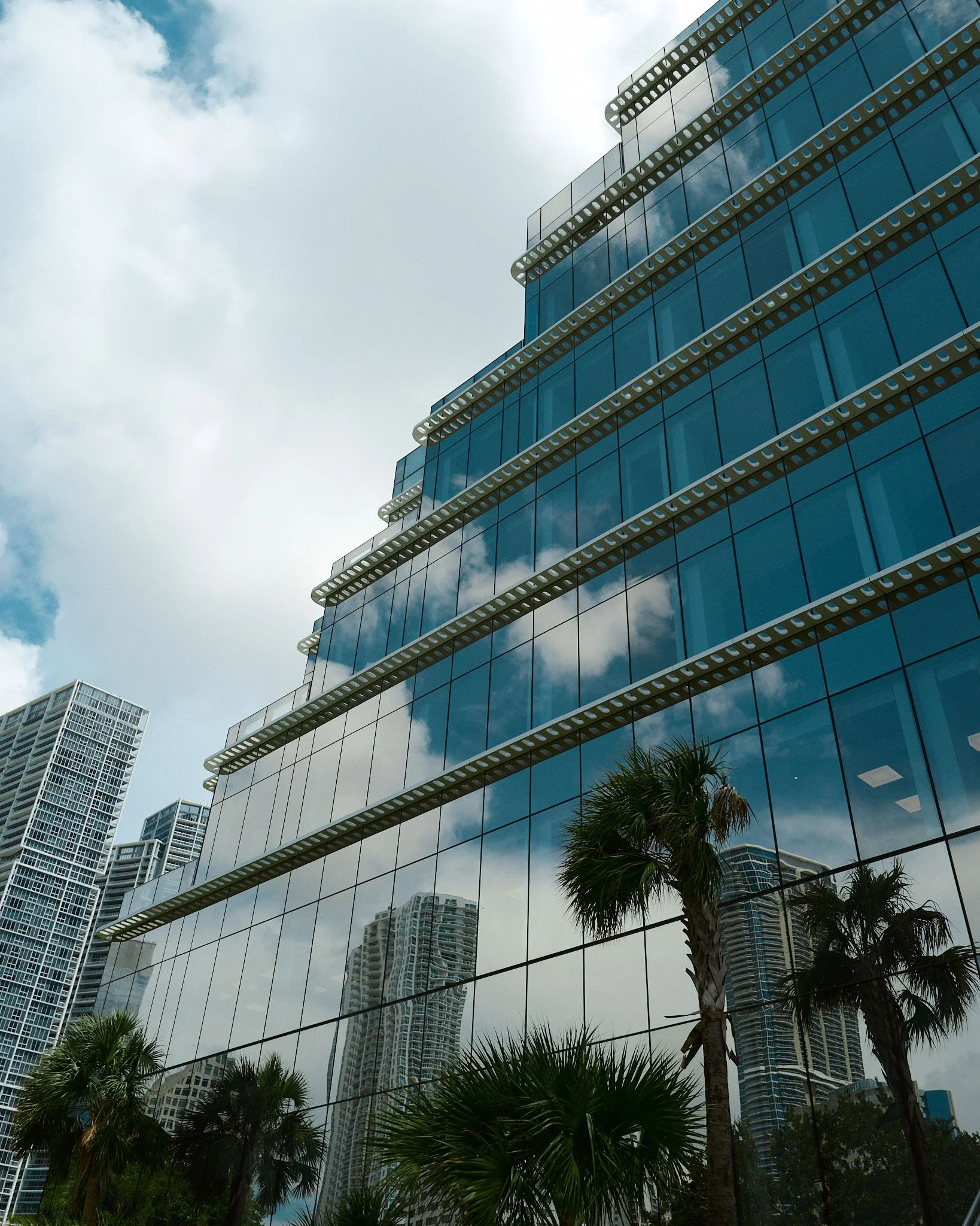Reflections of clouds and neighboring buildings in the glass windows of a modern high-rise building with palm trees in the foreground.
