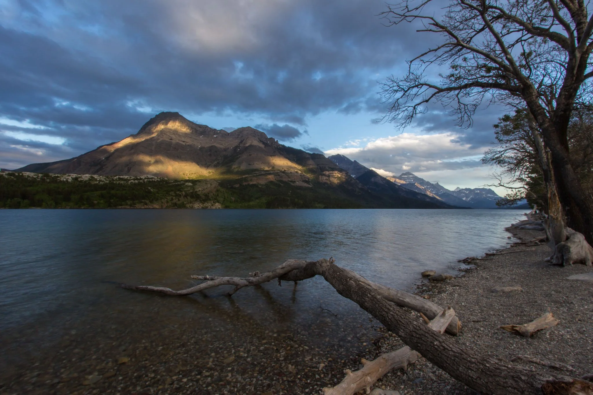A mountain landscape with a lake in the foreground, trees along the shore, and a cloudy sky overhead.