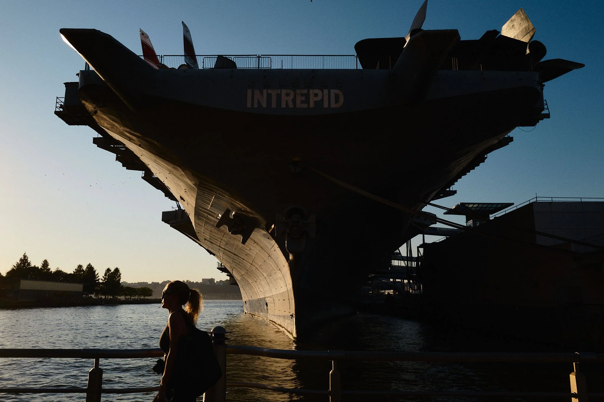 Silhouette of a woman standing by a railing near the USS Intrepid aircraft carrier, with the ship's bow and flight deck visible against a sunset sky.