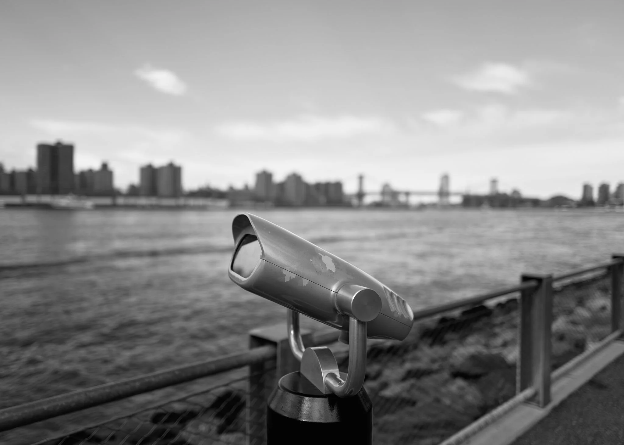 A coin-operated viewing telescope facing a river with a city skyline and a bridge in the distance, on a paved walkway near a metal railing.
