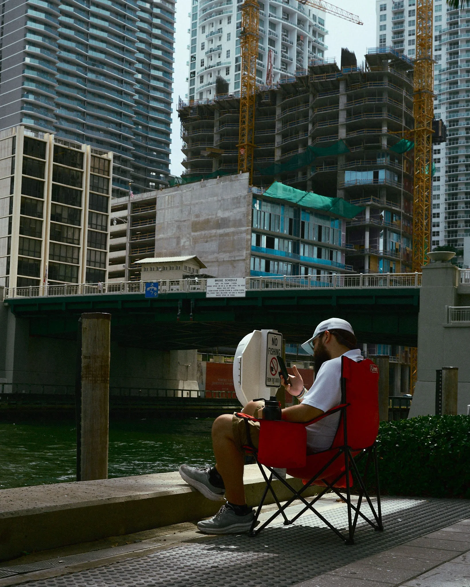 A man sitting on a red camping chair by the water, charging his phone next to a 