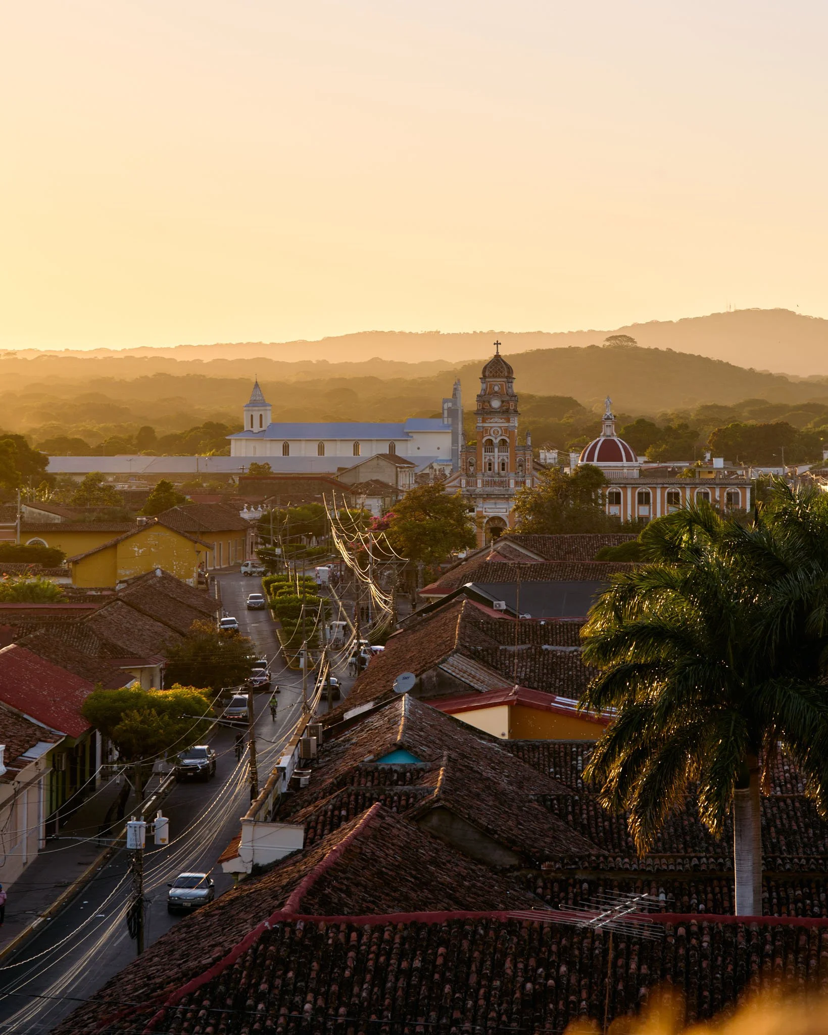 Sunset view of a small town with red-tiled roofs, a large palm tree, and a church with a dome and bell tower against a backdrop of hills.