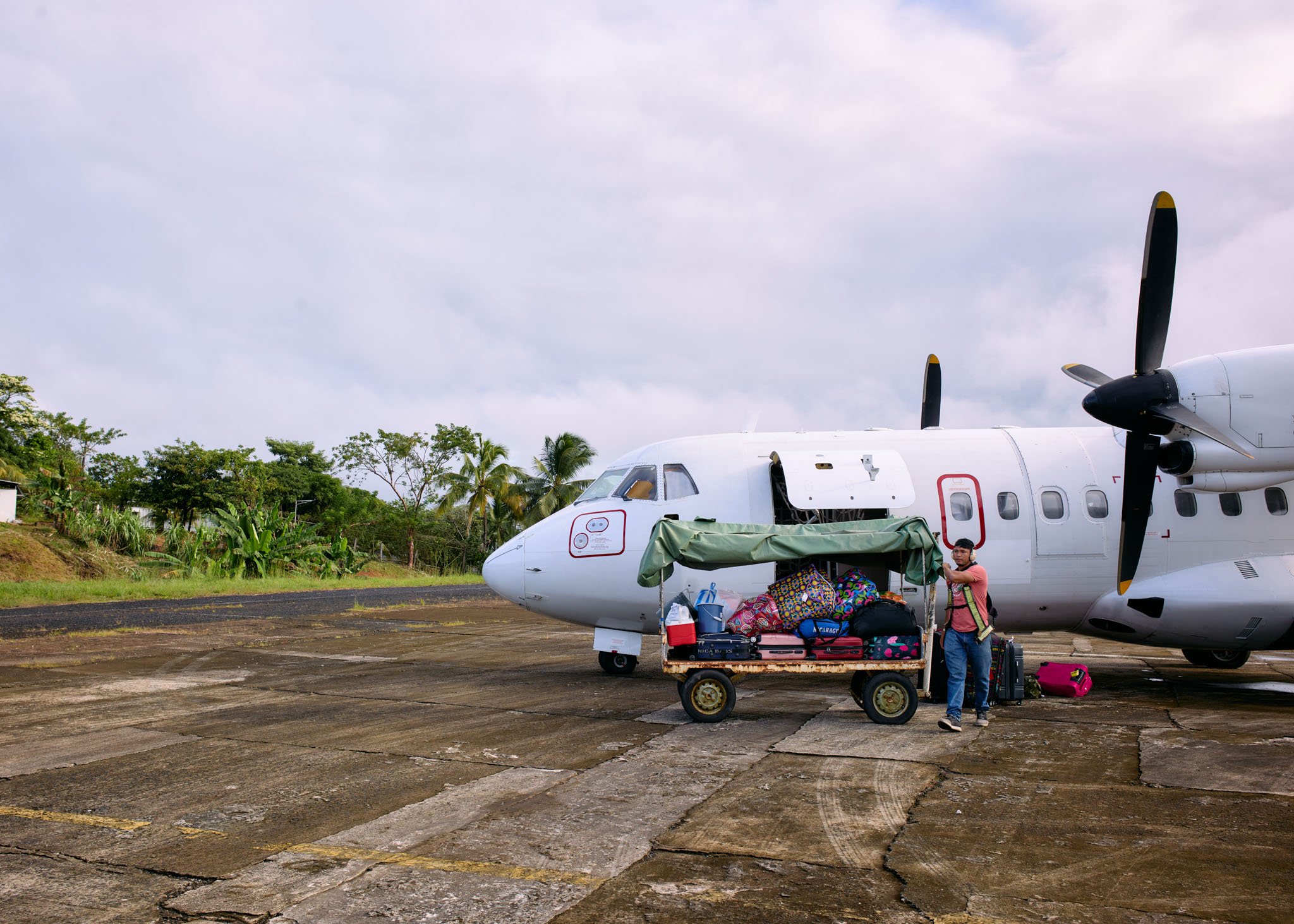 A small white commuter airplane parked on the tarmac with luggage and a red cart loaded with bags. A man is standing next to the cart, pulling a suitcase.