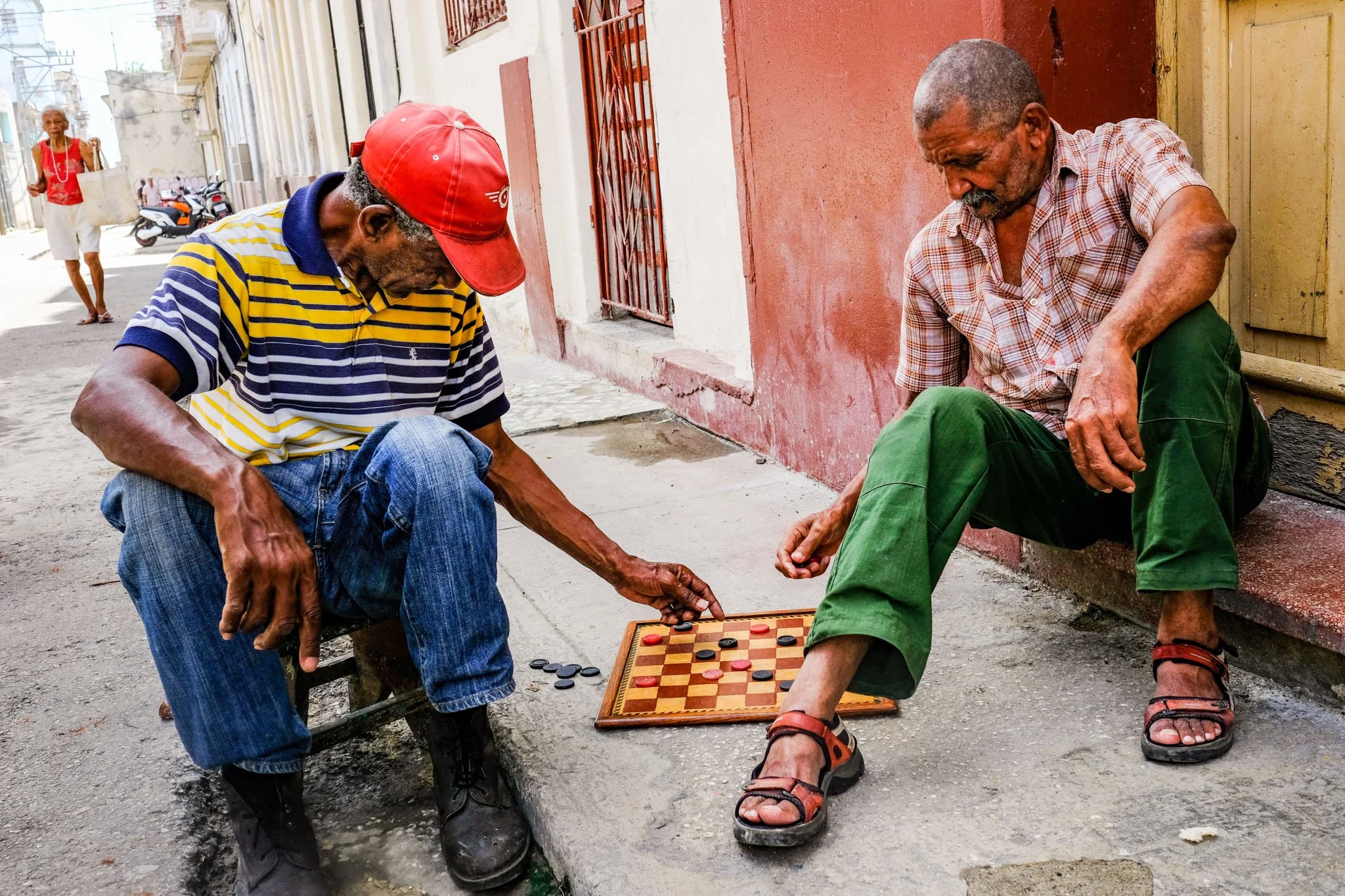 Two elderly men are playing checkers on a small wooden board on the sidewalk, with one man reaching for a piece. A woman is walking in the background on a sunny street lined with colorful buildings.