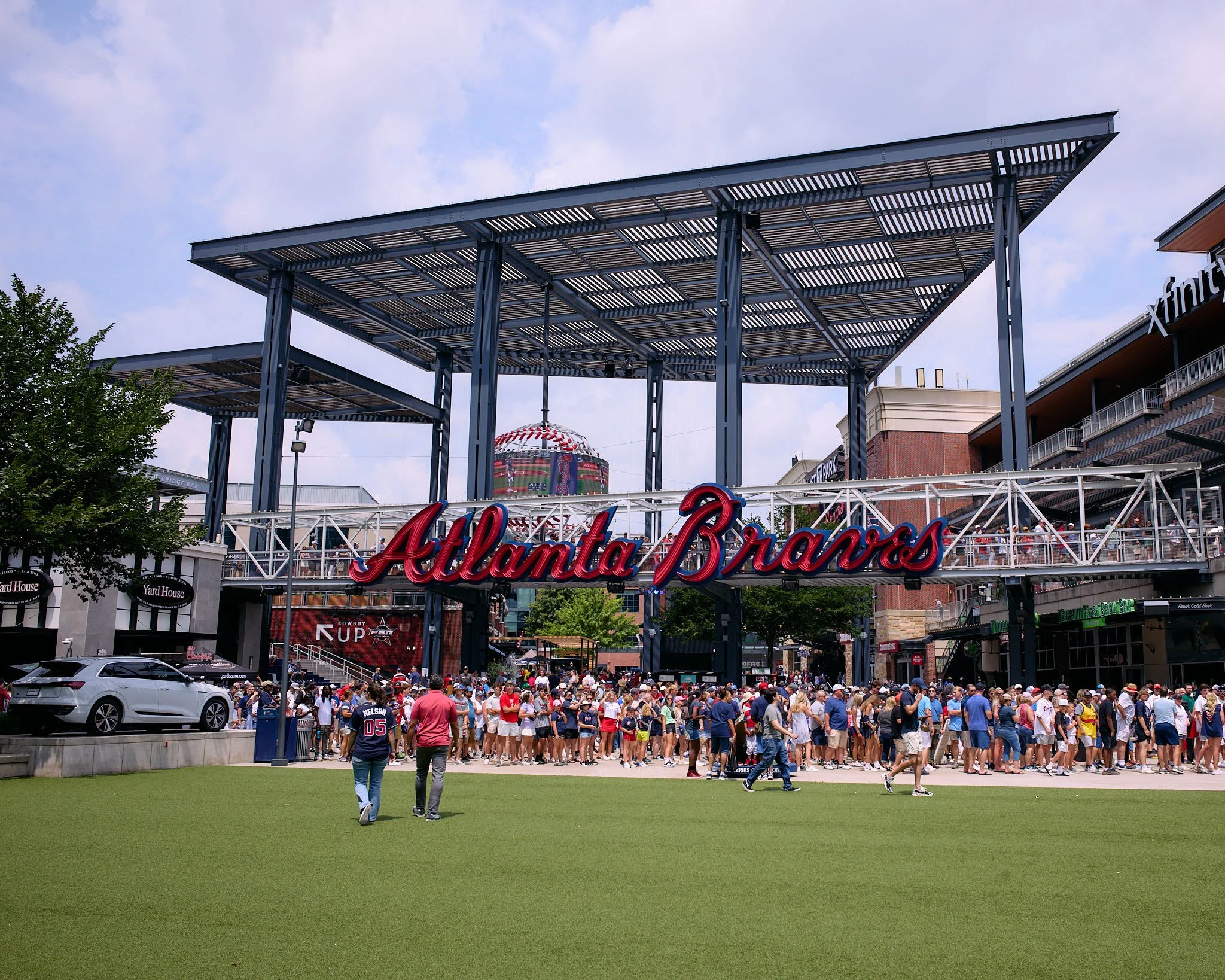 Entrance of Truist Park with a crowd of fans gathered outside, Atlanta Braves sign, and a partly cloudy sky.