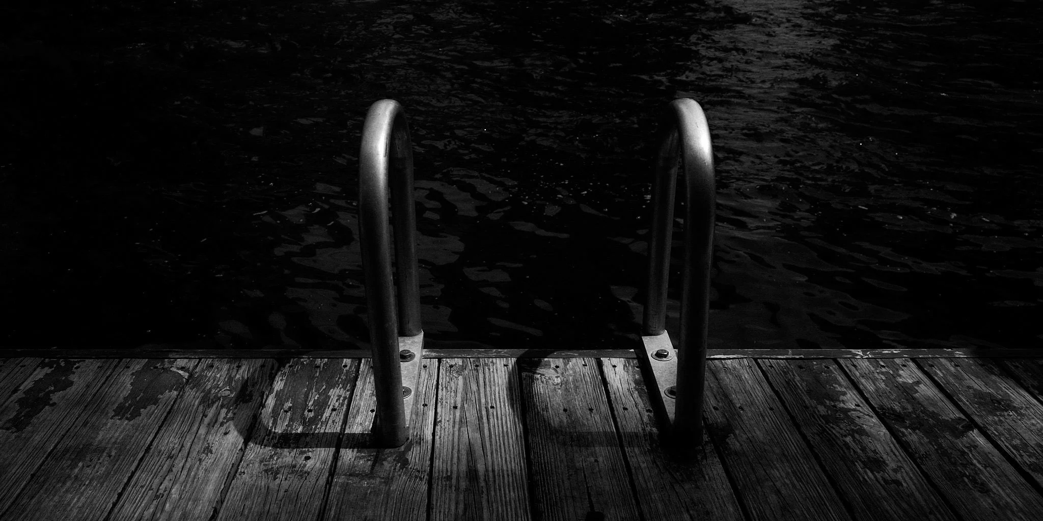 Black and white photo of a metal ladder leading into dark water, with a wooden dock in the foreground.