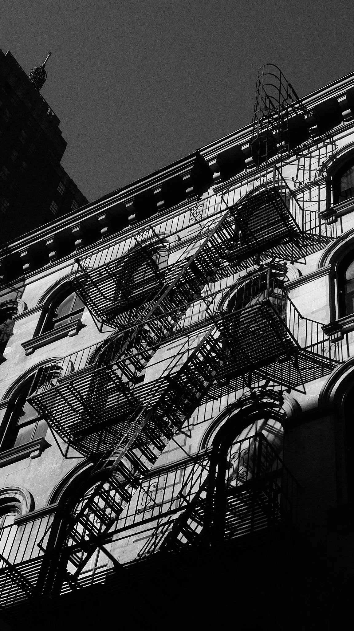 Black and white photo of a building with fire escapes on the exterior, with shadows cast on the facade.