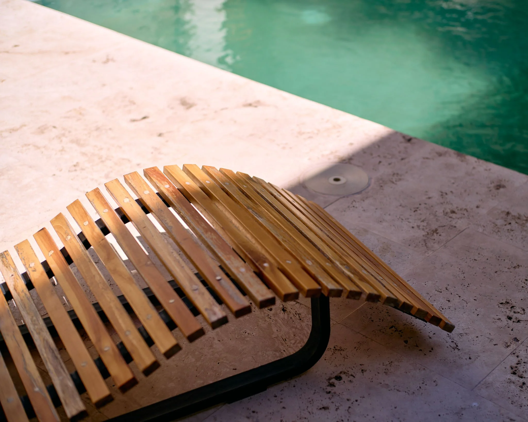 A wooden bench with a curved slatted design beside a swimming pool with turquoise water, on a stone deck.