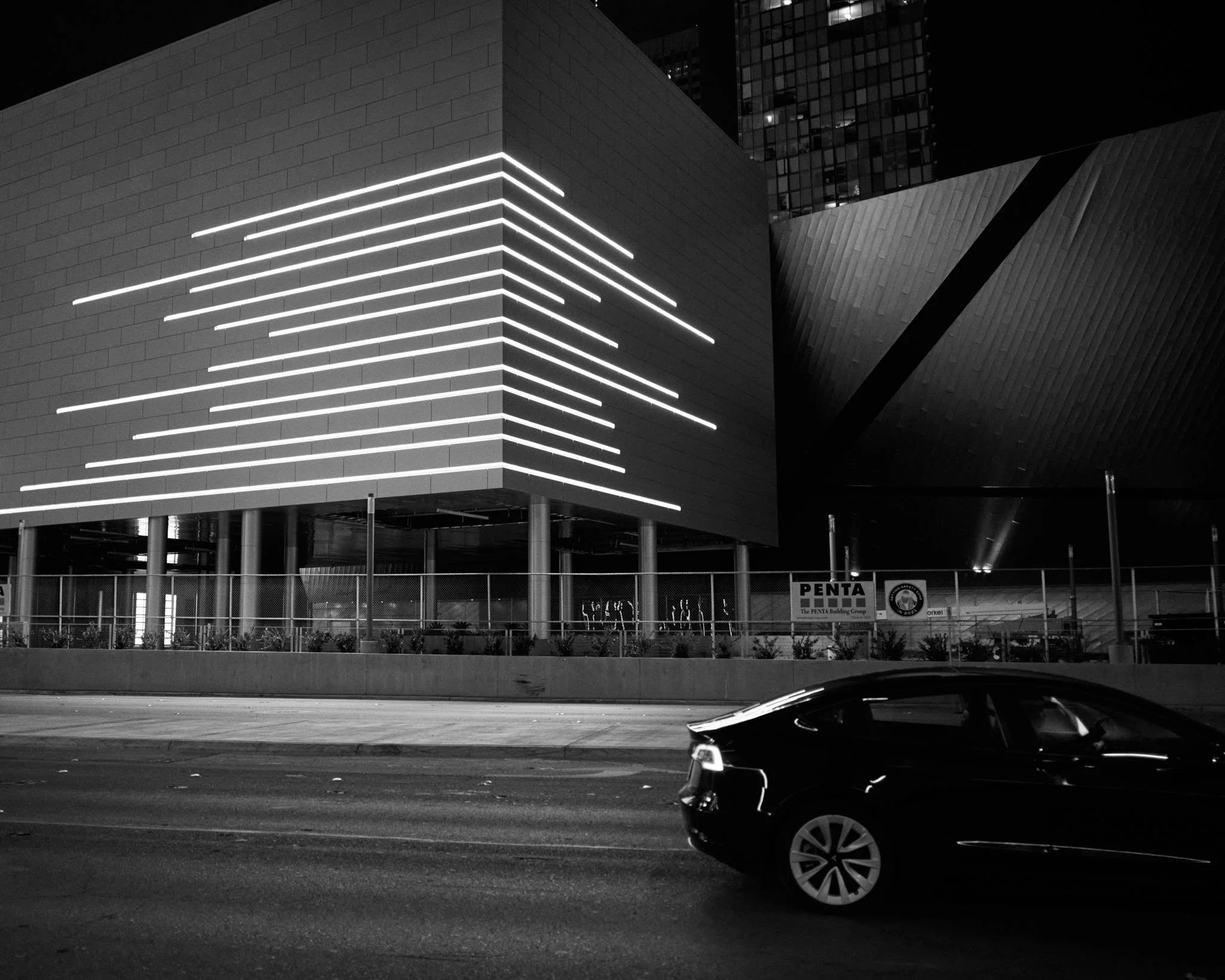 Night city scene with modern building featuring horizontal neon lights, a car on the road, and a construction fence with signs.