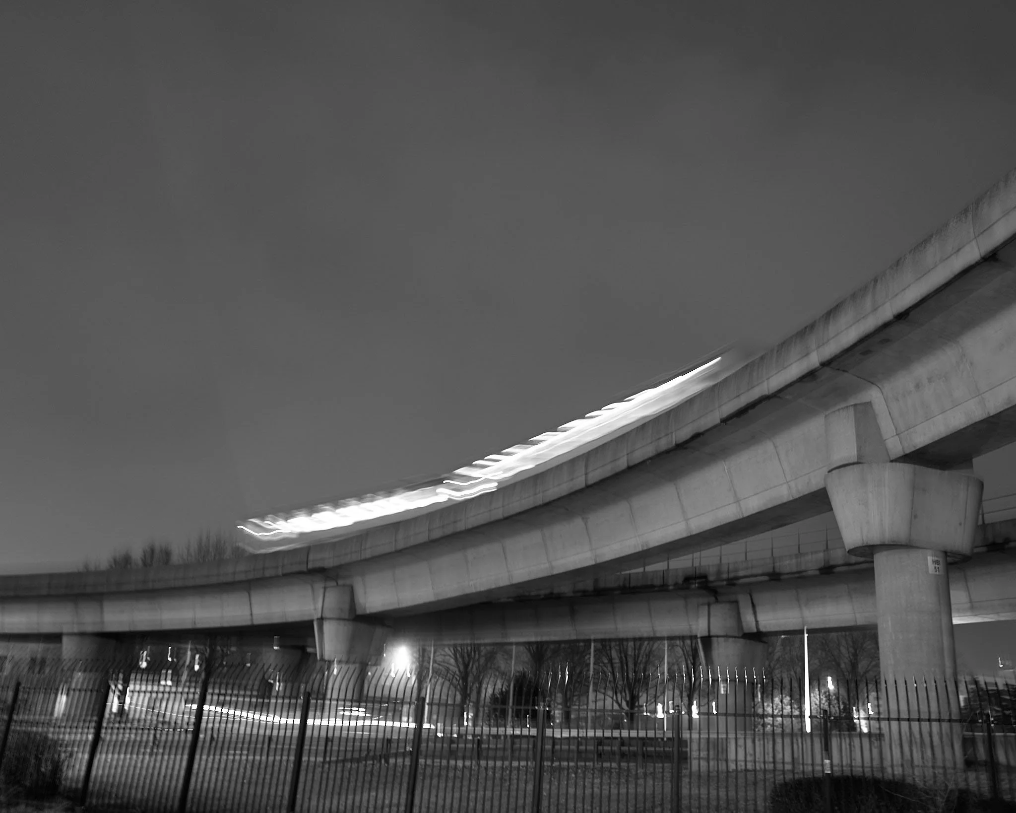 Black and white photo of an elevated highway at night with blurred motion of vehicles, a fence in the foreground, and trees and streetlights in the background.