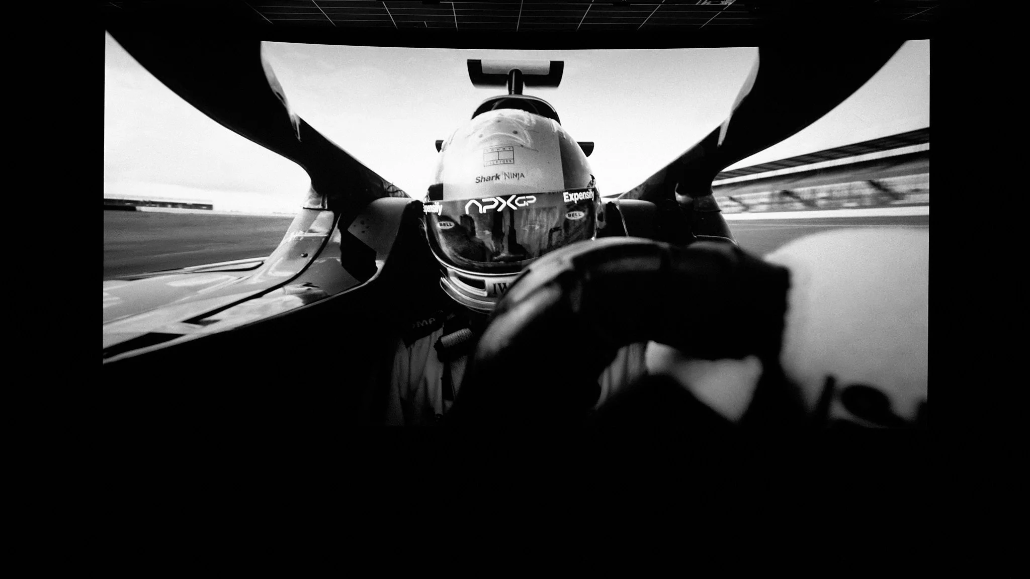 Black and white photo of a race car driver in a helmet, viewed through the windshield from inside the car, on a racetrack.