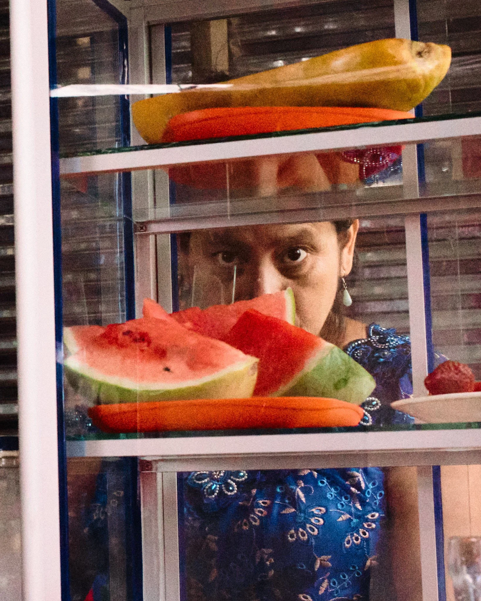 Person peeking through a glass display case with slices of watermelon, papaya, and other fruits inside.