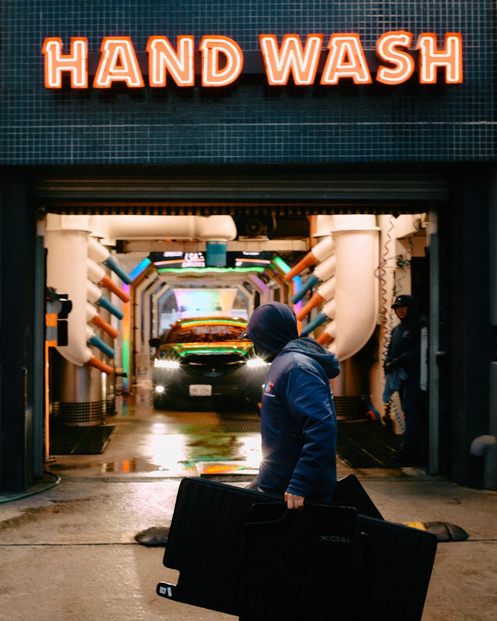 A person in a hooded jacket holding a guitar case walking out of a car wash tunnel with colorful pipes and lights. An employee wearing a cap and gloves is standing nearby.