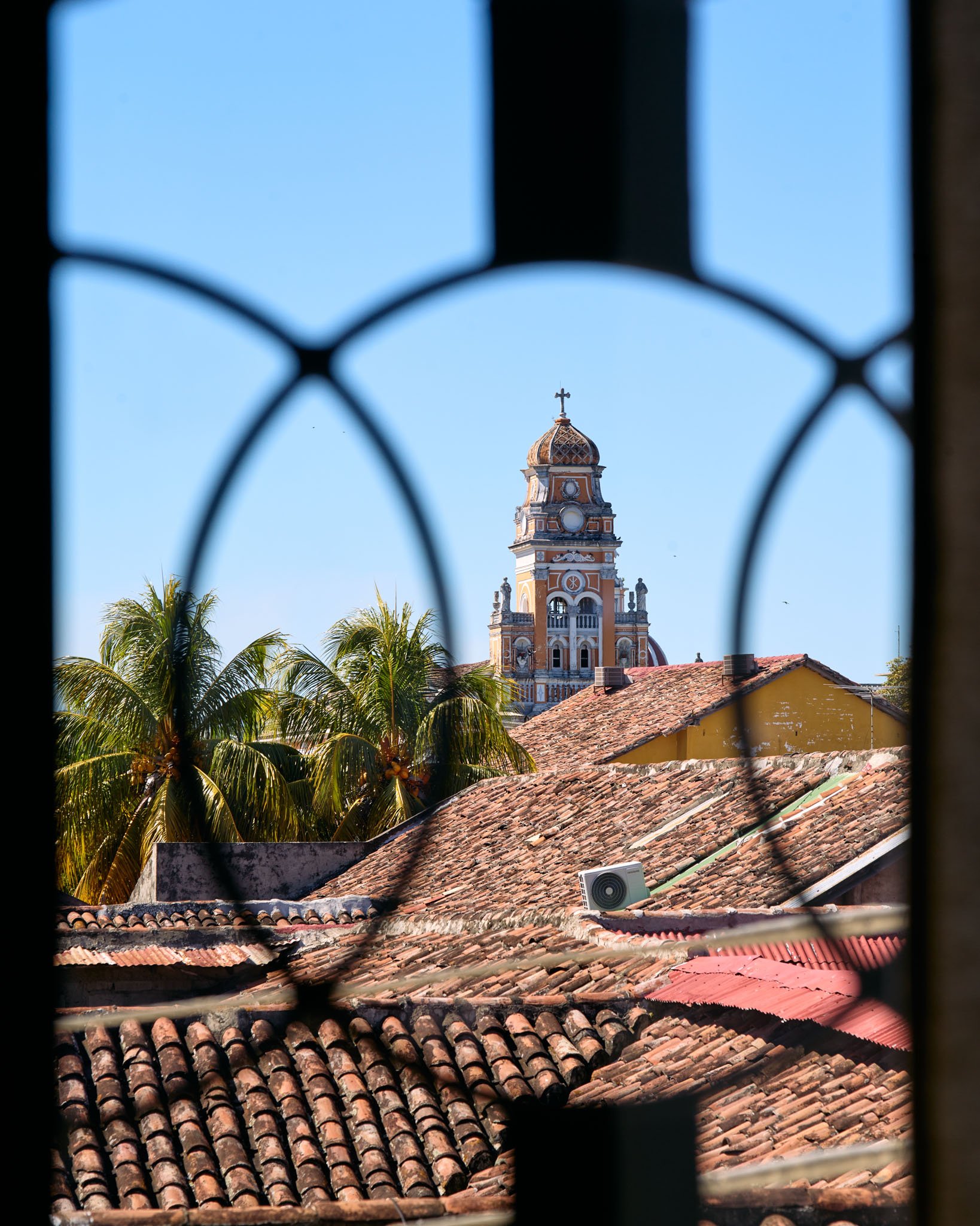View of a yellow church with a clock tower and cross on top, framed through a decorative iron window, with palm trees and tiled rooftops in the foreground under a clear blue sky.