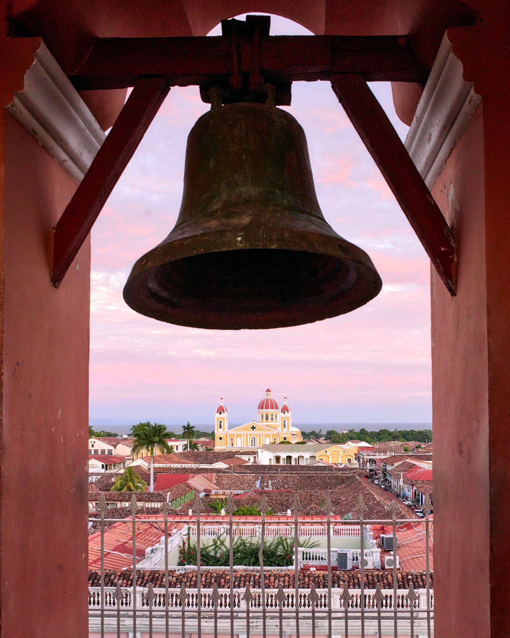 View of a church with yellow walls and red domes seen through a bell tower with a large metal bell hanging inside.